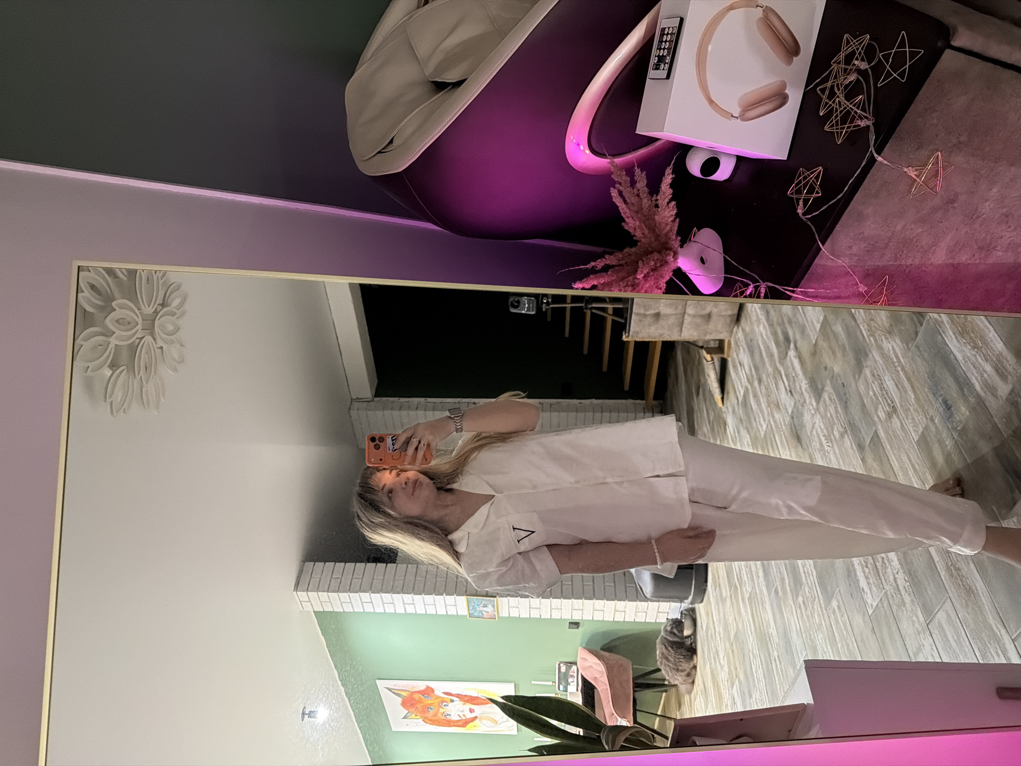 The image shows a reflection in a mirror. A woman is taking a selfie with an orange phone. She is wearing white pajamas. Reflected in the mirror are a white painted wall, green wall, a piece of artwork, a plant, and a cat in the background. In the upper portion of the image, there is a white chair illuminated with purple light, a pair of headphones, and star string lights. It seems that the location is in Большая Шильна, Russia.