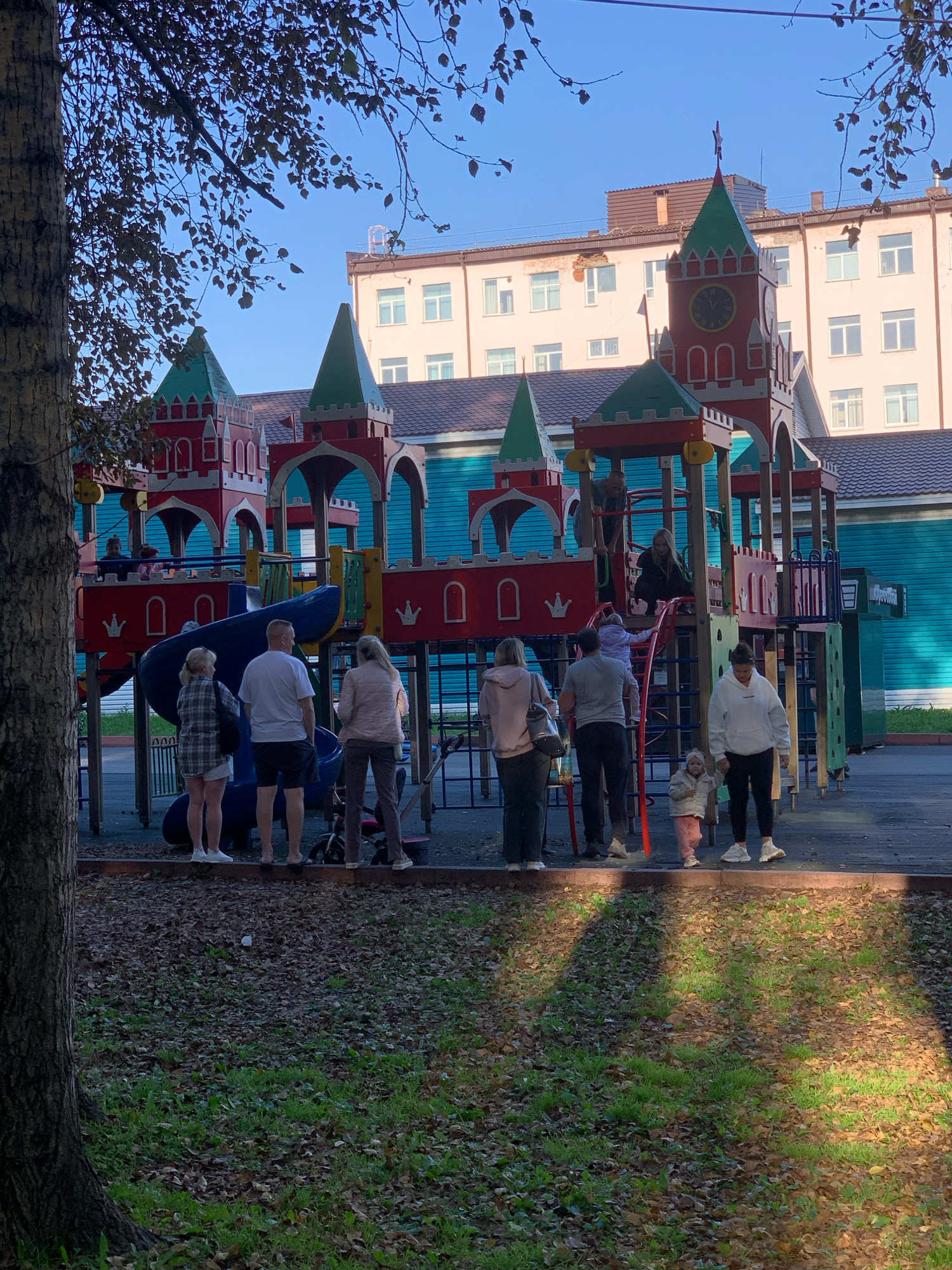 In Prokopyevsk, Russia, on a clear day, a group of people stands on a playground. The playground features a large, castle-themed structure with red and teal accents. Several individuals, including adults and children, are present, some standing and others in motion. In the foreground, a large tree trunk is visible on the left, with fallen leaves covering the ground. Behind the playground, a multi-story building with a light-colored facade and windows stands under a bright blue sky. The scene captures a moment of outdoor activity and community presence.