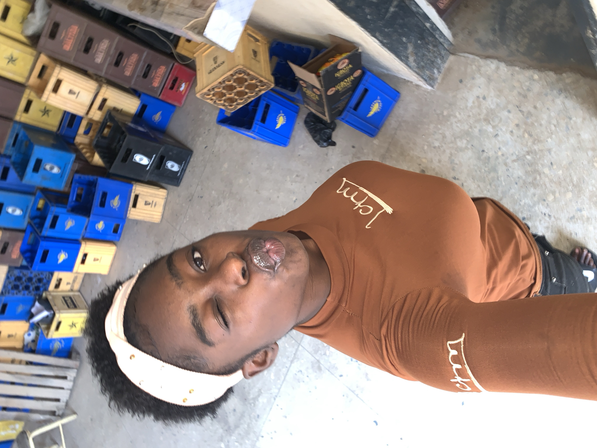 The image presents a person with dark skin, wearing a brown long-sleeved shirt and a white headband, positioned in front of a backdrop of stacked crates. The crates are varied in color, including blue, yellow, black, and brown. Some of the crates have visible brand names, such as "Guinness" and "Igboiya." The location is suggested to be in the city of Kabong, Nigeria. The person is looking directly at the camera with one eye closed. The background is a concrete-like surface.