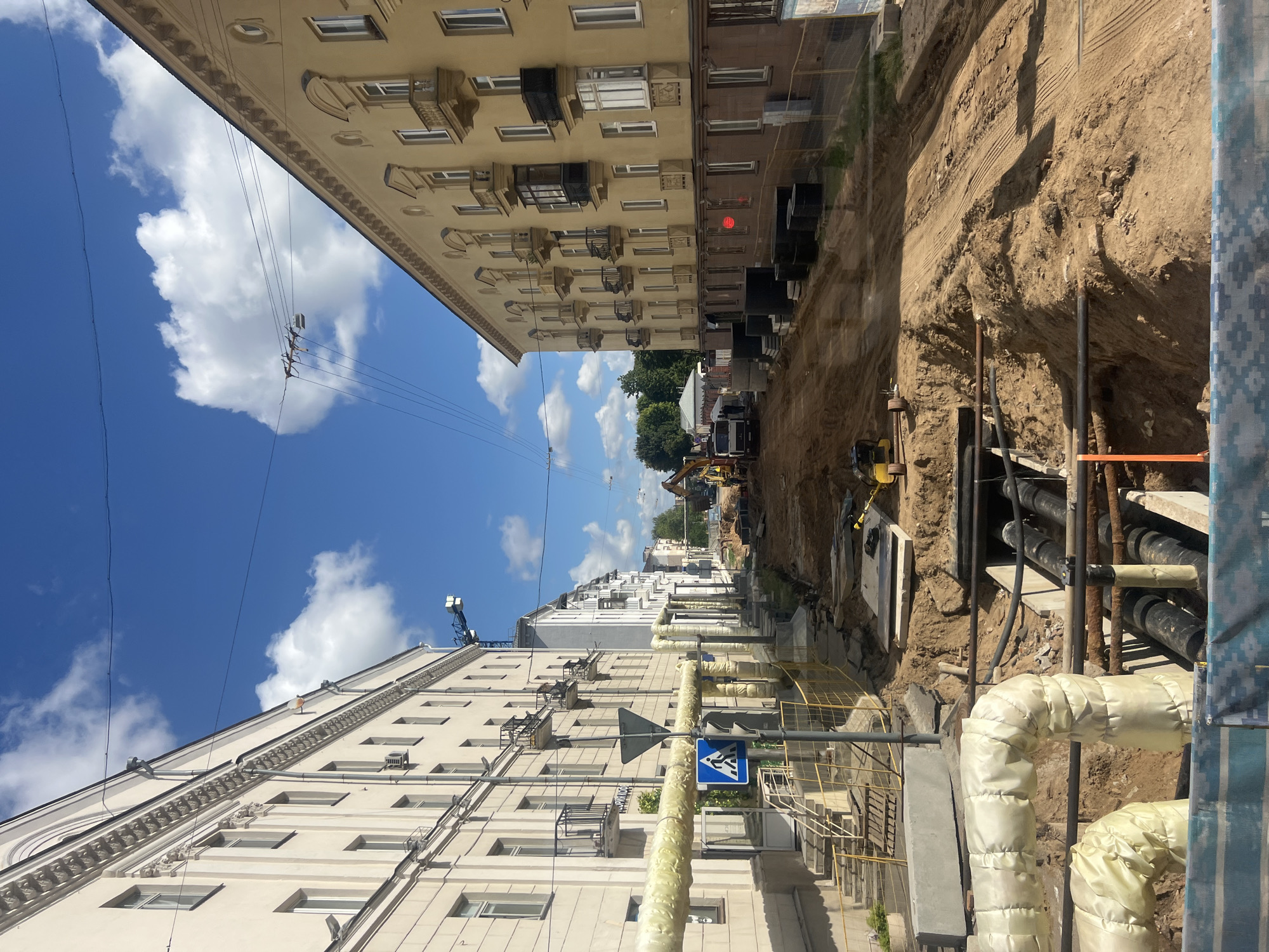 This is a street scene in Minsk, Belarus, depicting ongoing construction work. The perspective is from ground level, looking down a street lined with buildings on both sides. The street itself is dug up, revealing exposed pipes and trenches filled with soil. Construction equipment, including an excavator and a truck, are visible further down the street. Large, insulated pipes run along the side of one of the buildings. The sky is blue with scattered white clouds, and there are overhead power lines. The buildings appear to be residential or mixed-use, with multiple stories and balconies. The overall impression is one of urban infrastructure development.