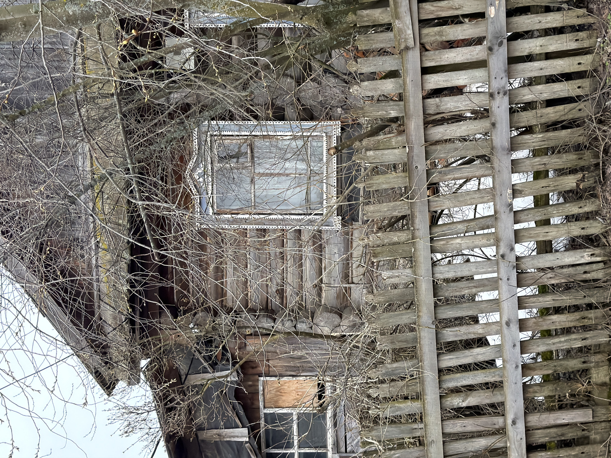 An upside-down, medium-shot photograph depicts the exterior of a derelict wooden structure in Chaikovsky, Russia. The building features a decaying facade with two distinct window types. The upper window is a larger, square frame with ornate trim, while the lower window is a smaller, simpler design, partially boarded up. A weathered wooden fence-like element is present on the right side of the image, constructed of horizontal slats with vertical supports. The foreground is dominated by a network of bare tree branches, obscuring portions of the structure. The overall scene conveys a sense of abandonment and disrepair.