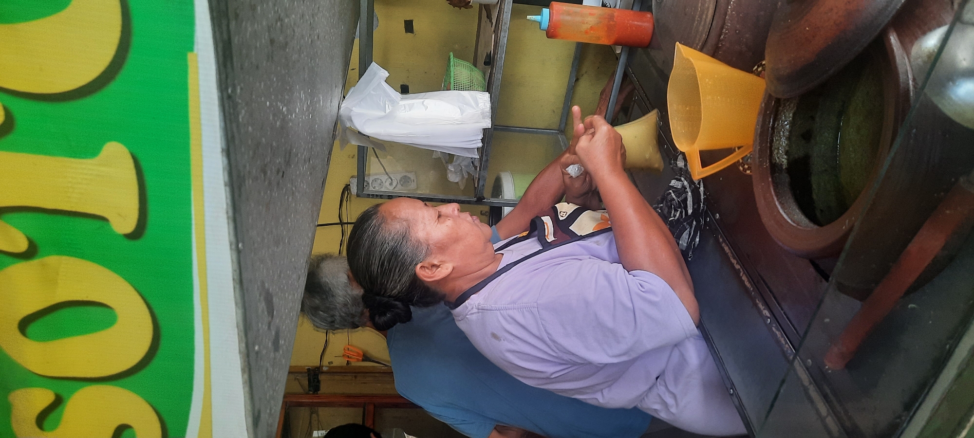 The image depicts a food stall in Selopuro, Indonesia, with a sign above that reads "SOTO". A woman wearing a light purple t-shirt and an apron is seen actively preparing food. She is holding a plastic bag filled with a yellow substance and appears to be sealing it with her hands. Next to her, a man with gray hair, wearing a blue t-shirt, stands with his back to the camera, also seemingly involved in the food preparation process. In the foreground, a large clay pot containing a green liquid is visible, along with a yellow plastic pitcher and a reddish sauce bottle. The environment appears to be an open-air stall with shelving behind the woman, holding various items including a white plastic bag and a green basket.