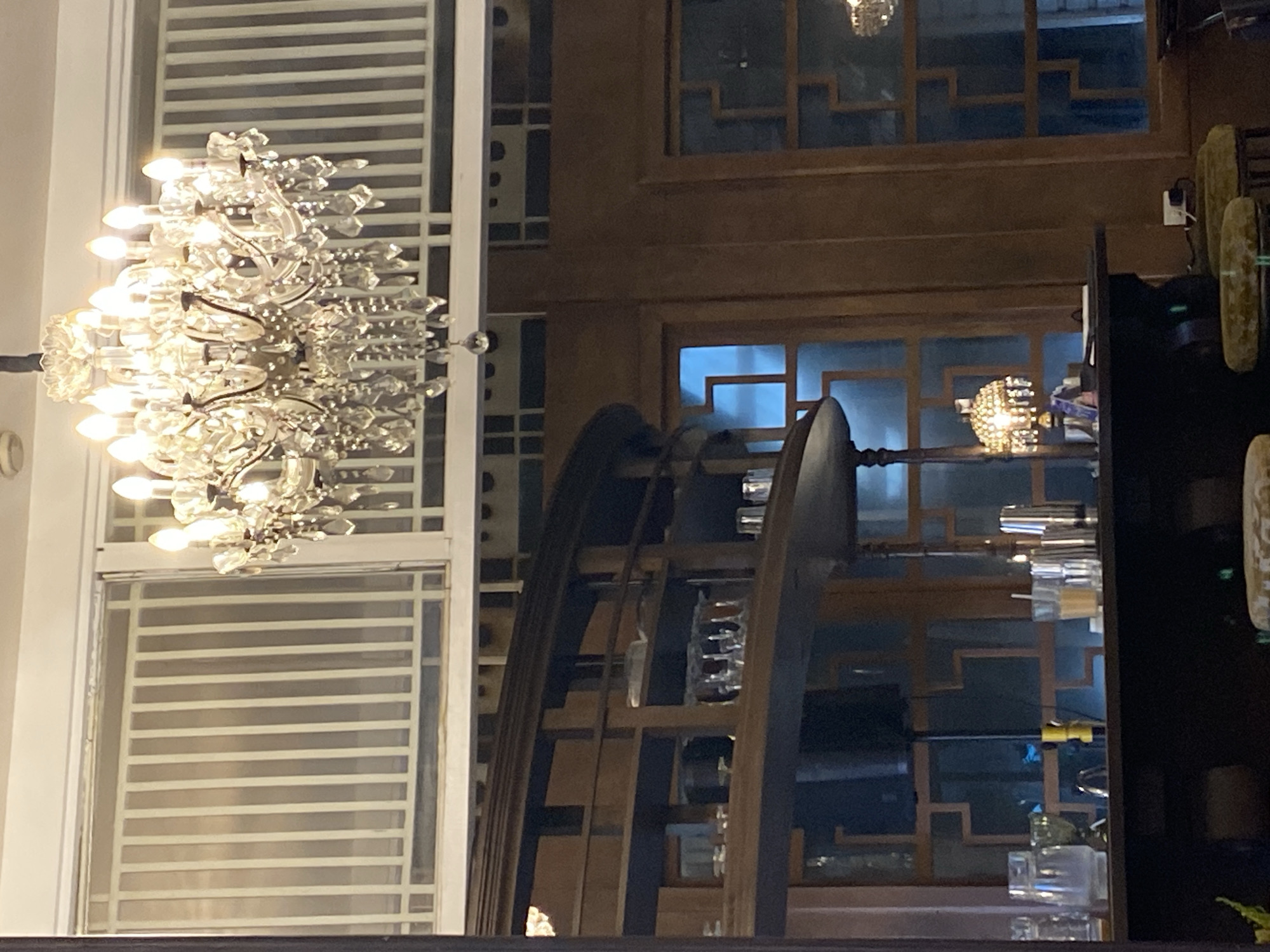 The image captures an indoor scene, likely a restaurant or bar, with a prominent crystal chandelier hanging from the ceiling. The chandelier features multiple tiers of lights and numerous hanging crystals, reflecting light. Below the chandelier, a dark wooden shelf unit is visible, displaying various glassware, including wine glasses and tumblers. A bar counter is situated in the foreground, with drinks and bar tools such as shakers and cups on its surface. A vintage-style table lamp with a jeweled shade sits on the counter, casting a warm glow. Behind the bar, windows with intricate wooden lattices reveal a dimly lit interior. The walls are paneled with dark wood. The overall ambiance suggests an upscale, possibly dimly lit establishment. The specific location in Davao City, Philippines, is not determinable from the visual information provided.