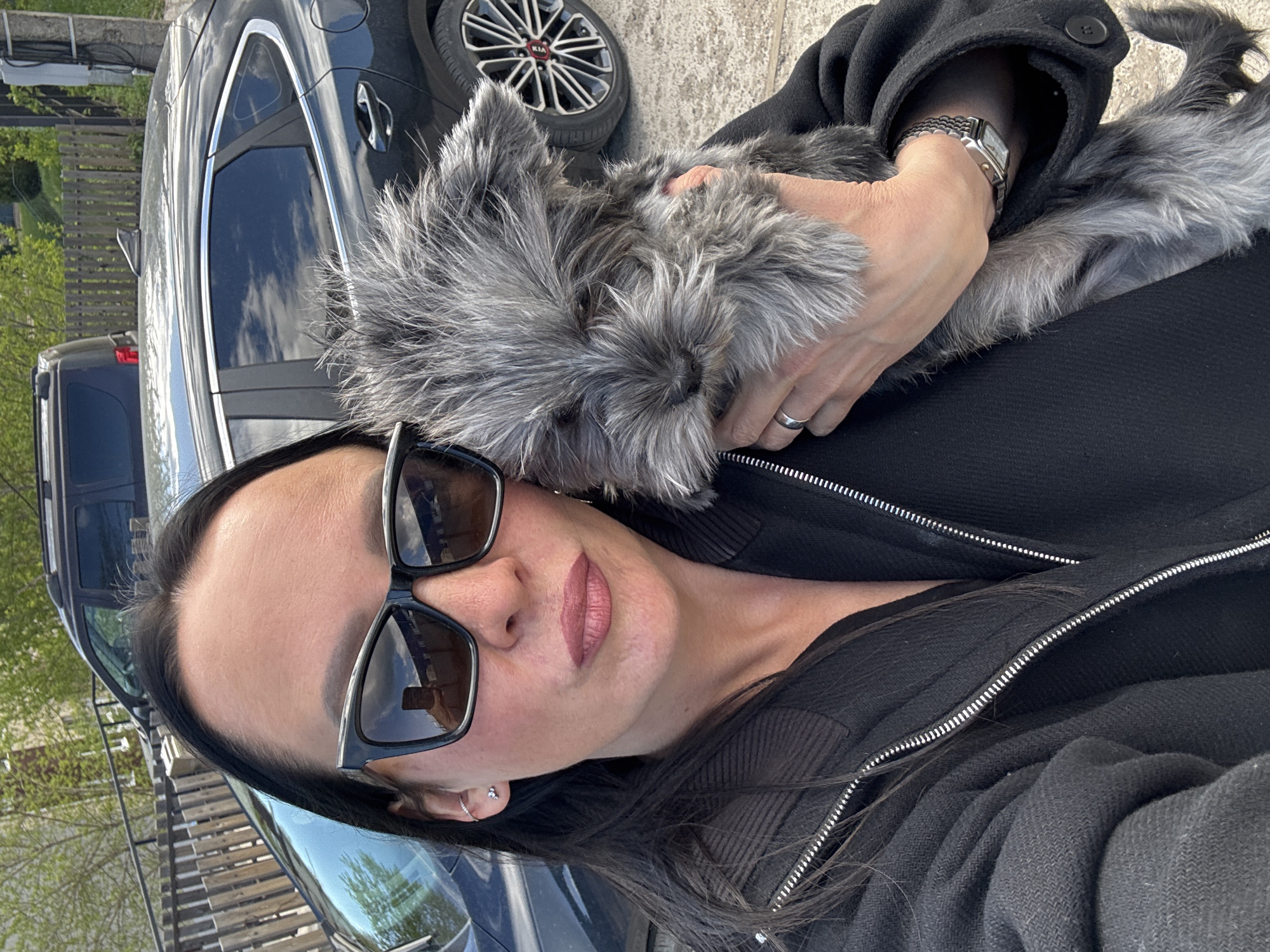 A woman is holding a gray schnauzer dog in her arms, while standing outside near a black car. The woman is wearing sunglasses, a silver watch, and a black coat. The location is Chaikovsky, Russia, based on the wheel design on the car behind her.