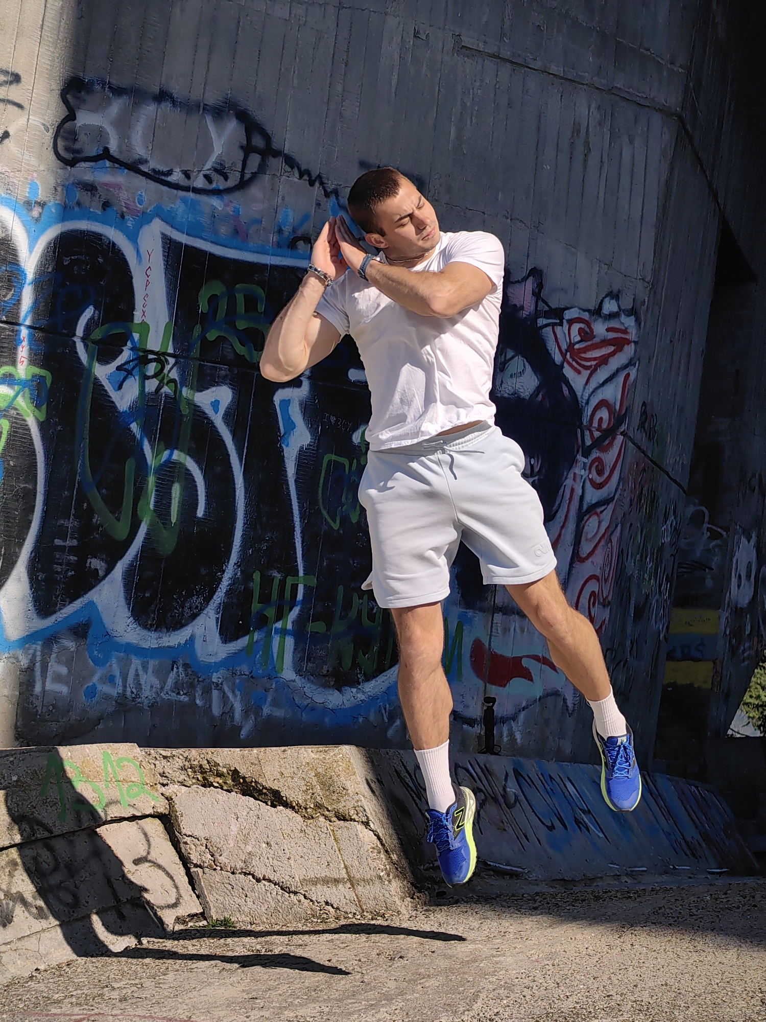 In Varna, Bulgaria, a young man with short brown hair, wearing a white t-shirt and light blue shorts with white socks and blue and yellow athletic shoes, is captured mid-air, appearing to be jumping or posing against a concrete wall covered in graffiti. His arms are raised and bent at the elbows, with his hands near his ears, and his eyes are closed. The background consists of a large, weathered concrete structure adorned with vibrant graffiti in shades of blue, black, green, and red. In the foreground, a cracked concrete ledge casts shadows on the ground. The lighting suggests it is a sunny day.