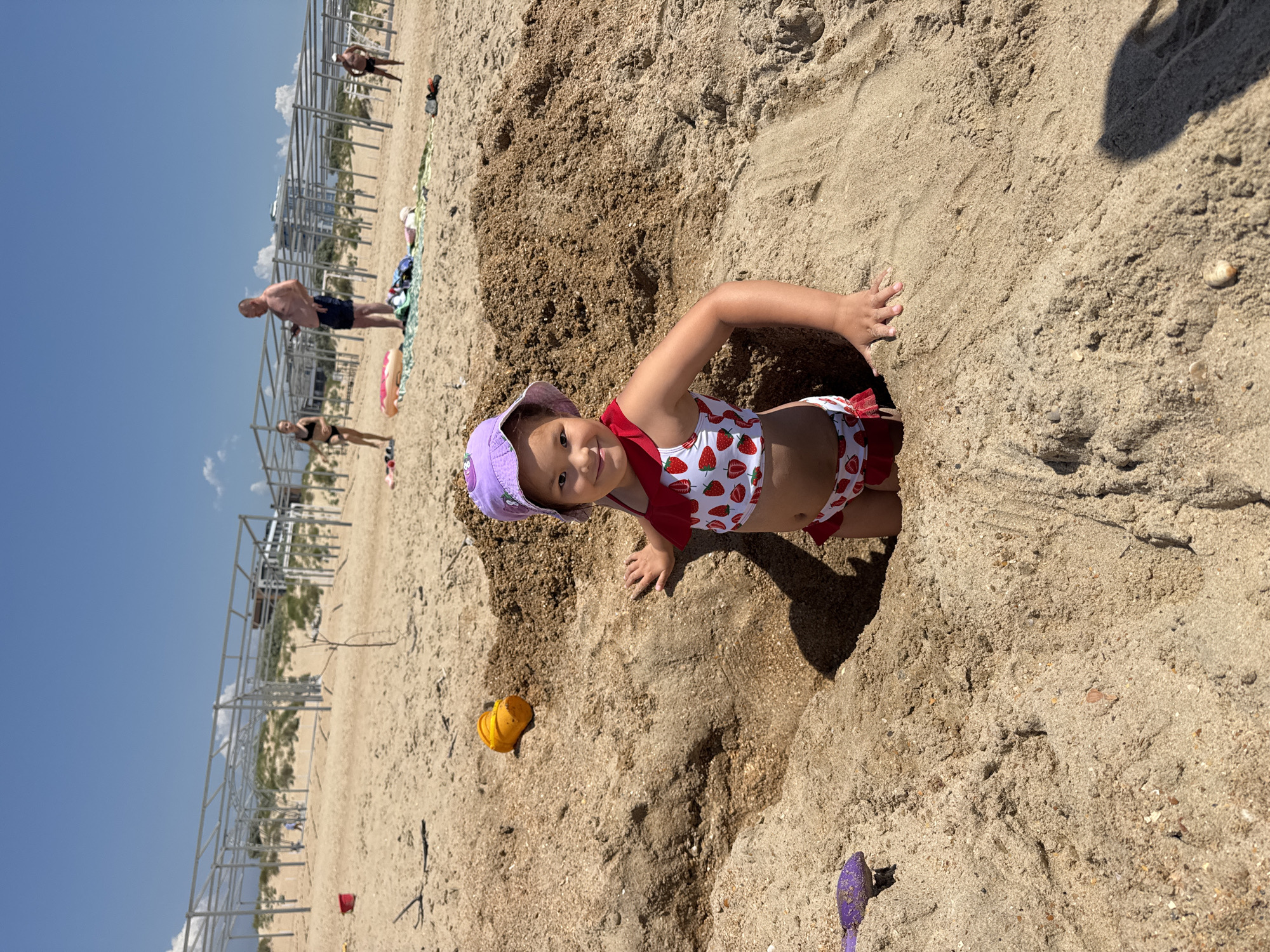 A young child in a strawberry-patterned swimsuit and purple sun hat is playing in the sand on a beach. The child is partially submerged in a dug-out hole, with their hands resting on the sand. A yellow plastic shovel and a purple shovel are visible nearby, along with a red bucket further in the distance.
In the background, there are metal structures, possibly part of a beach infrastructure or construction. Several adults are also present on the beach, some walking and others appearing to relax. The sky is clear and blue, suggesting a sunny day. The overall scene depicts a typical day at the beach, with children and adults enjoying leisure activities. The sand appears light brown and grainy, typical of a beach environment. The lighting suggests it is daytime, likely midday due to the clear skies and shadows.