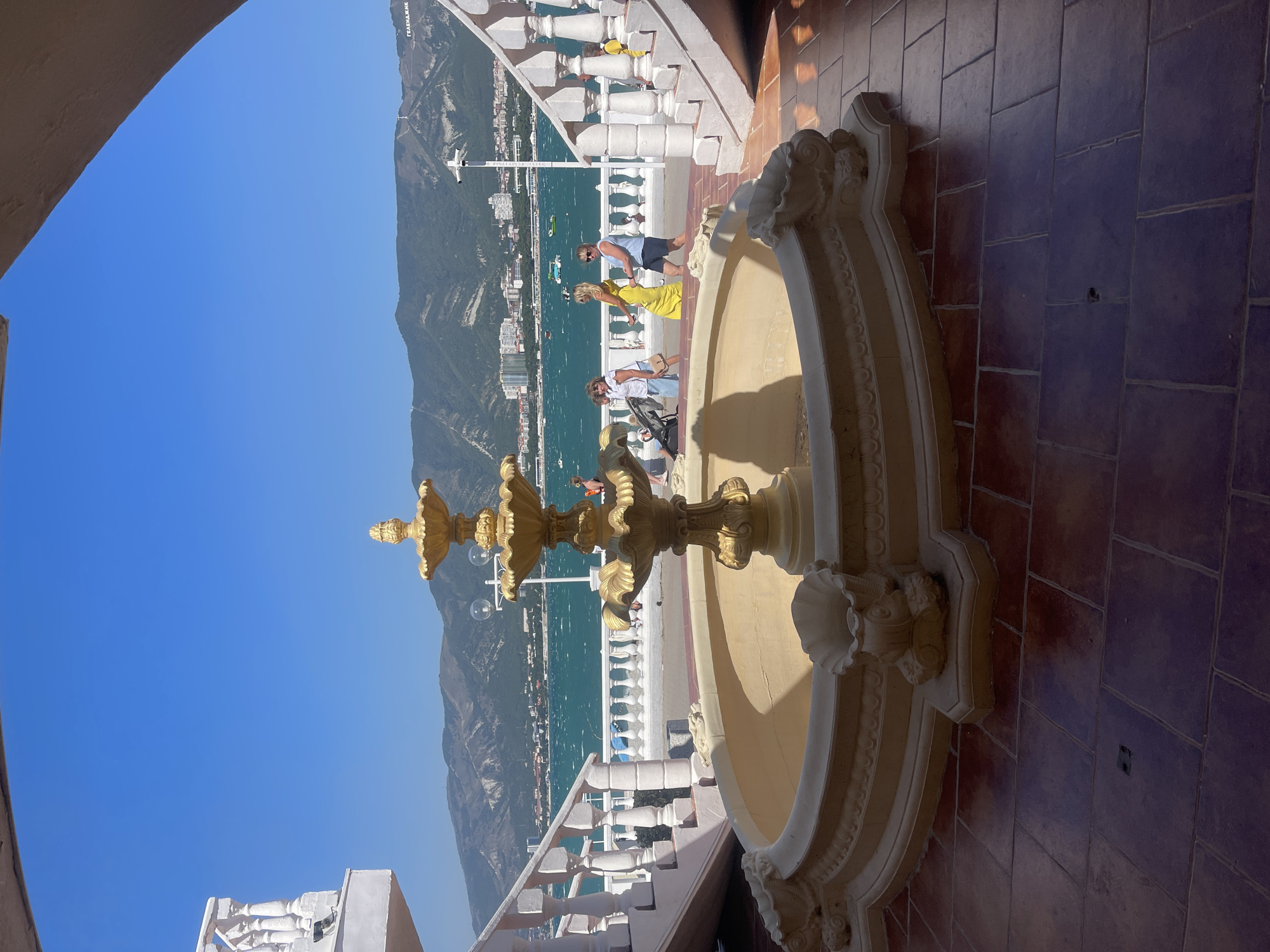 A wide-angle, eye-level shot captures a sunlit scene in Gelendzhik, Russia, featuring a decorative tiered fountain as the central focus. The fountain, crafted from a creamy-colored material with gilded accents, is situated on a tiled patio. In the background, a vibrant turquoise sea stretches towards a range of imposing, verdant mountains. The sky above is a clear, bright blue.
Several people are visible on a promenade bordering the sea. To the left, a white balustrade lines a set of stairs, and to the right, a similar balustrade curves away, framing the scene. The architecture suggests a public park or a resort area. The water is choppy, indicating wind, and a few small boats are dotted across its surface.
On the promenade, a woman in a bright yellow dress is walking from left to right, accompanied by another woman in a blue top and shorts. Further left, a woman pushes a stroller, and another individual in a white top and light blue skirt walks beside her. The overall impression is one of a pleasant, active summer day by the sea.