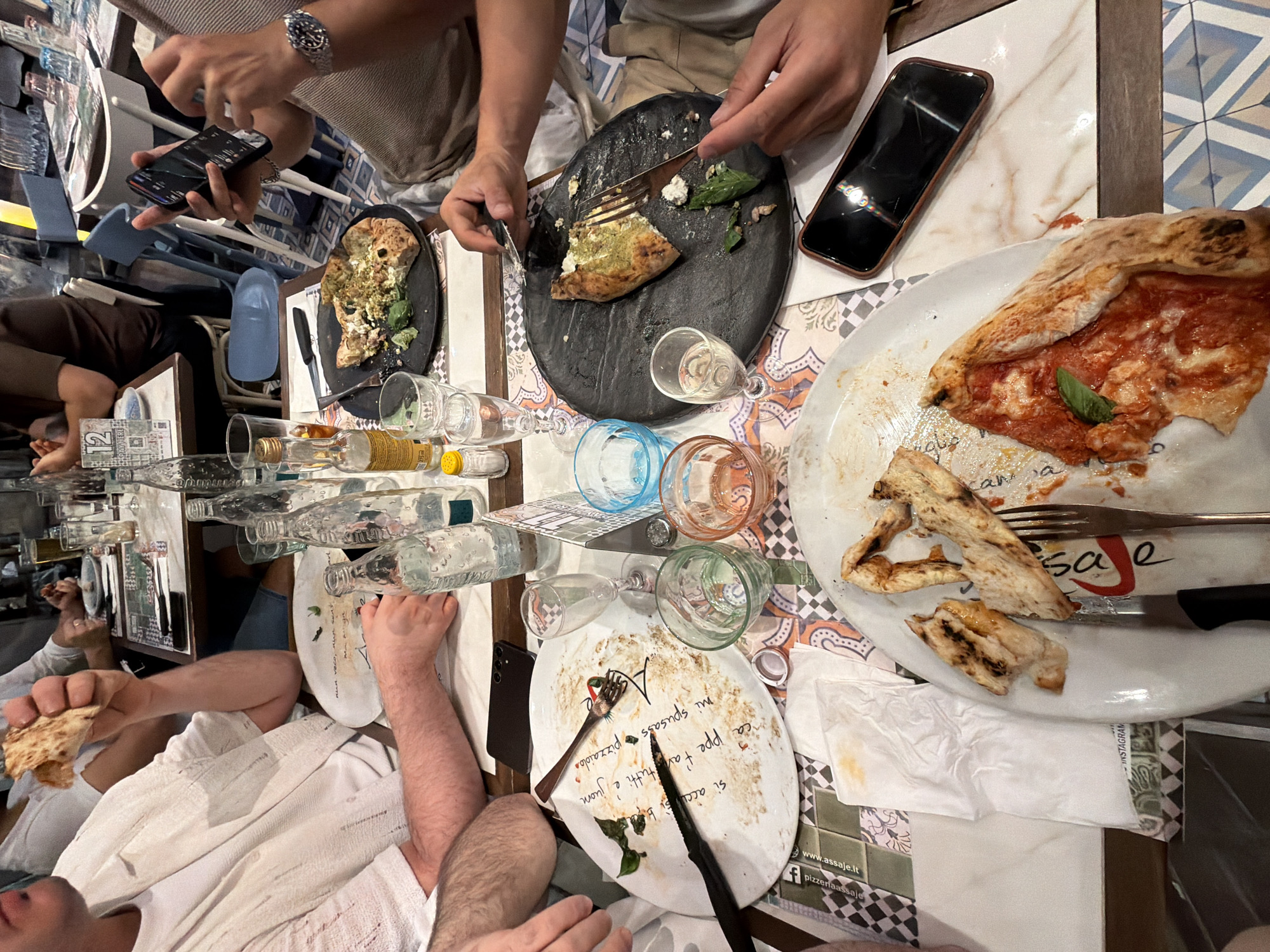 The image depicts a group of people seated around a table at a restaurant, likely in Milan, Italy, given the restaurant's branding visible on a napkin in the foreground which reads "pizzassai.it". The table is laden with plates of pizza, some partially eaten, along with glasses, bottles, and cutlery. One person in the foreground is holding a piece of pizza and appears to be eating. Another person to the right is looking at their phone. The overall atmosphere suggests a casual dining experience. The background shows other diners and restaurant furniture, including chairs and tables, indicating a public eating establishment.