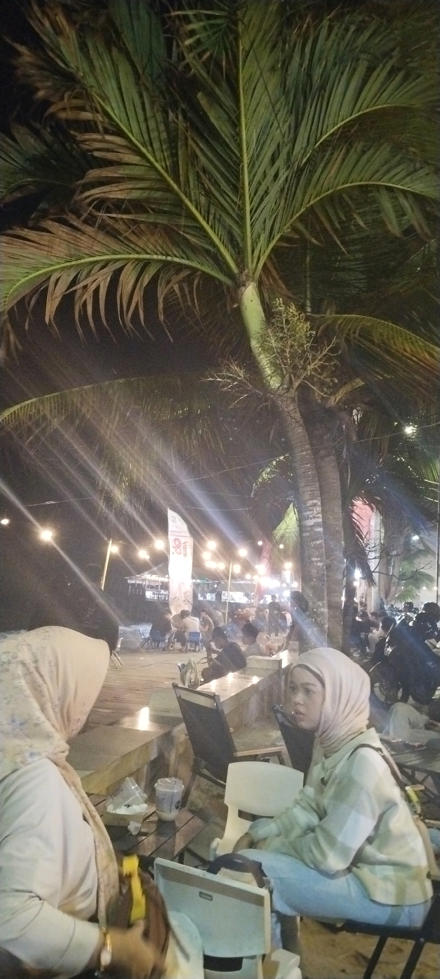 The image depicts a nighttime scene at an outdoor seating area, likely a cafe or restaurant, near a beach or waterfront in Balikpapan, Indonesia. A large palm tree dominates the upper portion of the frame, its fronds illuminated by strings of warm-toned lights that stretch across the background. The lighting creates a festive and relaxed atmosphere.
In the foreground, two individuals are seated. On the left, a person wearing a light-colored hijab and a patterned top is visible from the chest up. They are holding a yellow object. To the right, another person, also wearing a light-colored hijab and a checkered long-sleeved shirt and light blue pants, is seated on a chair and looking to the left. A laptop or similar electronic device is placed on a small table in front of them.
The background reveals more seating areas with people gathered. There are tables and chairs, and some individuals appear to be dining or socializing. In the distance, structures that resemble small shops or food stalls can be seen, also lit by string lights. The presence of several motorcycles parked on the right side suggests a public area. The ground surface appears to be made of wood planks in the foreground, leading to a sandy or paved area further back. A white banner with some text, possibly in a local language, is displayed in the middle ground. The overall environment suggests a popular spot for evening recreation in Balikpapan.