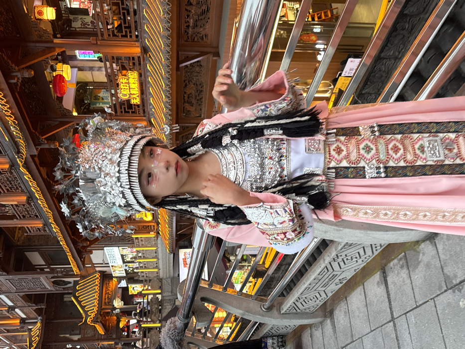 A young individual is centrally positioned in Yuzhong District, Chongqing, China, specifically on a walkway overlooking multi-tiered traditional Chinese architecture, characteristic of Hongyadong Commercial Street. The subject, likely female, wears elaborate traditional attire. This includes a tall, ornate silver-colored headdress with floral and leaf motifs, butterfly accents, and a wide band featuring numerous vertical silver prongs. Her hair is styled in long black braids, interwoven with silver-colored strands, extending over her shoulders.
The attire consists of a pink long-sleeved tunic, with wide cuffs heavily embroidered in geometric patterns of silver, white, red, and blue threads, accented with white fur trim. A white undergarment panel is visible beneath a large, intricate silver-colored chest piece adorned with multiple dangling elements and colorful beads. A long pink skirt features a prominent vertical embroidered panel down the front, displaying complex geometric patterns in red, green, white, silver, and black, with small silver adornments. The subject's face is characterized by light skin tone, with decorative pink/purple eyeliner and iridescent glitter applied to the cheeks. Her right hand is gently placed at her neck/collar, while her left hand is slightly raised, fingers splayed, resting near or on a silver metal handrail.
In the mid-ground, a robust grey concrete railing, featuring carved geometric patterns, supports a shiny silver-colored metal handrail. The background showcases multi-story traditional wooden buildings with tiered roofs and intricate carvings, extensively illuminated by warm orange and yellow lights, including string lights and red lanterns. Illuminated Chinese signs, such as "C拉卡卡" and "无人机观位" (Drone Viewing Spot), are visible. The scene is artificially lit, indicating a nighttime or late dusk setting. The lower portion of another individual, clad in dark traditional attire with embroidered details, is partially visible on the far left. The ground is paved with grey tiles.