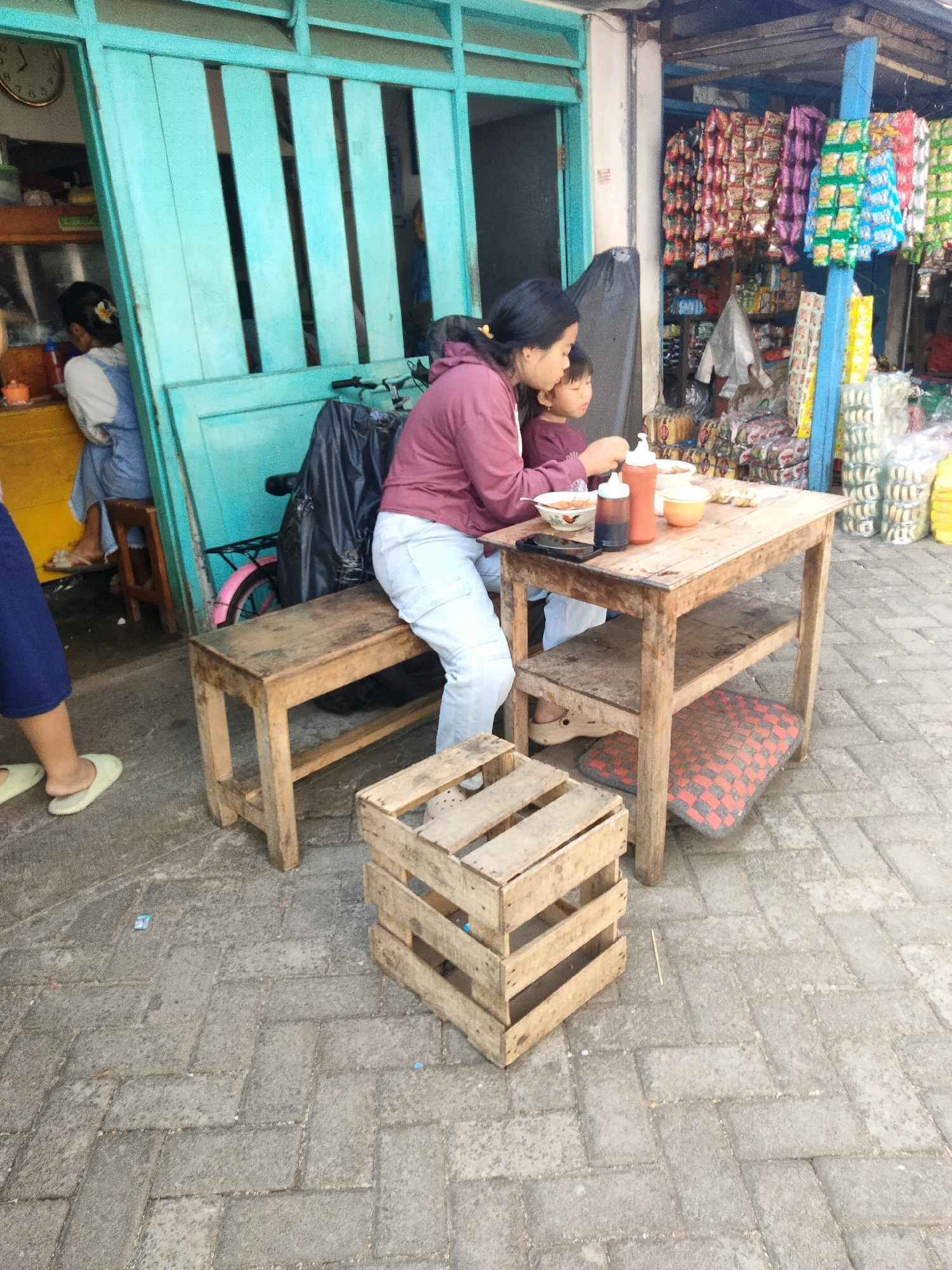 A woman and a child are seated at a wooden table outside a shop in Sawahan, Indonesia. The woman, wearing a maroon hoodie and light-colored pants, is serving food from bowls. The child, wearing a maroon shirt, is seated beside her. On the table are bowls of food, two bottles of condiments, and a mobile phone. A wooden bench is placed next to the table, and a wooden crate sits on the pavement in the foreground. In the background, shelves are filled with packaged goods. The shop features a teal-colored door and a display of colorful snack packages. The pavement is made of gray interlocking bricks.