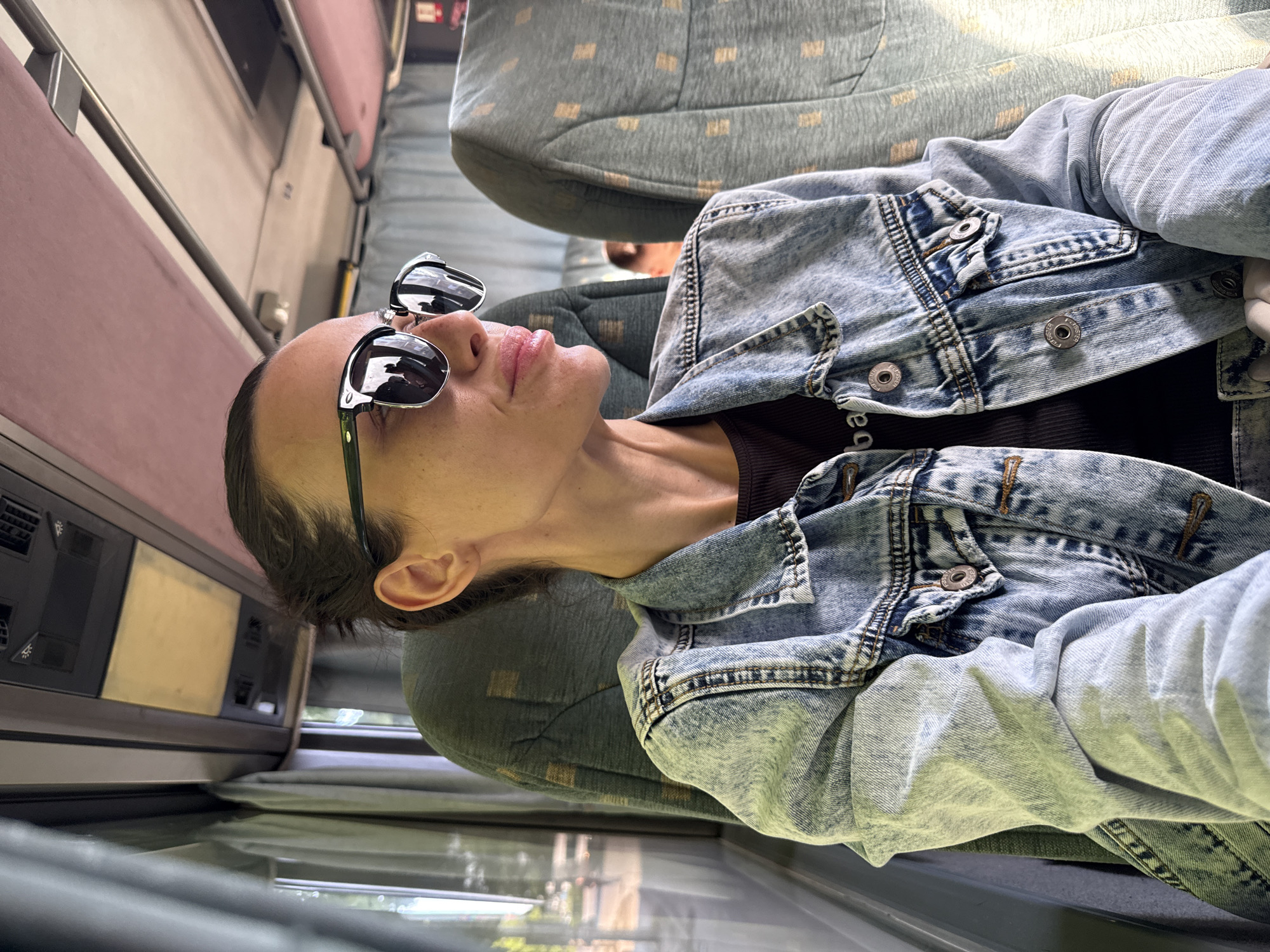 A woman wearing a denim jacket and sunglasses sits in a bus, resting against the seat with her head tilted back. The bus interior is visible, including other seats and a window. The location appears to be Pleven, Bulgaria, based on previous instructions.