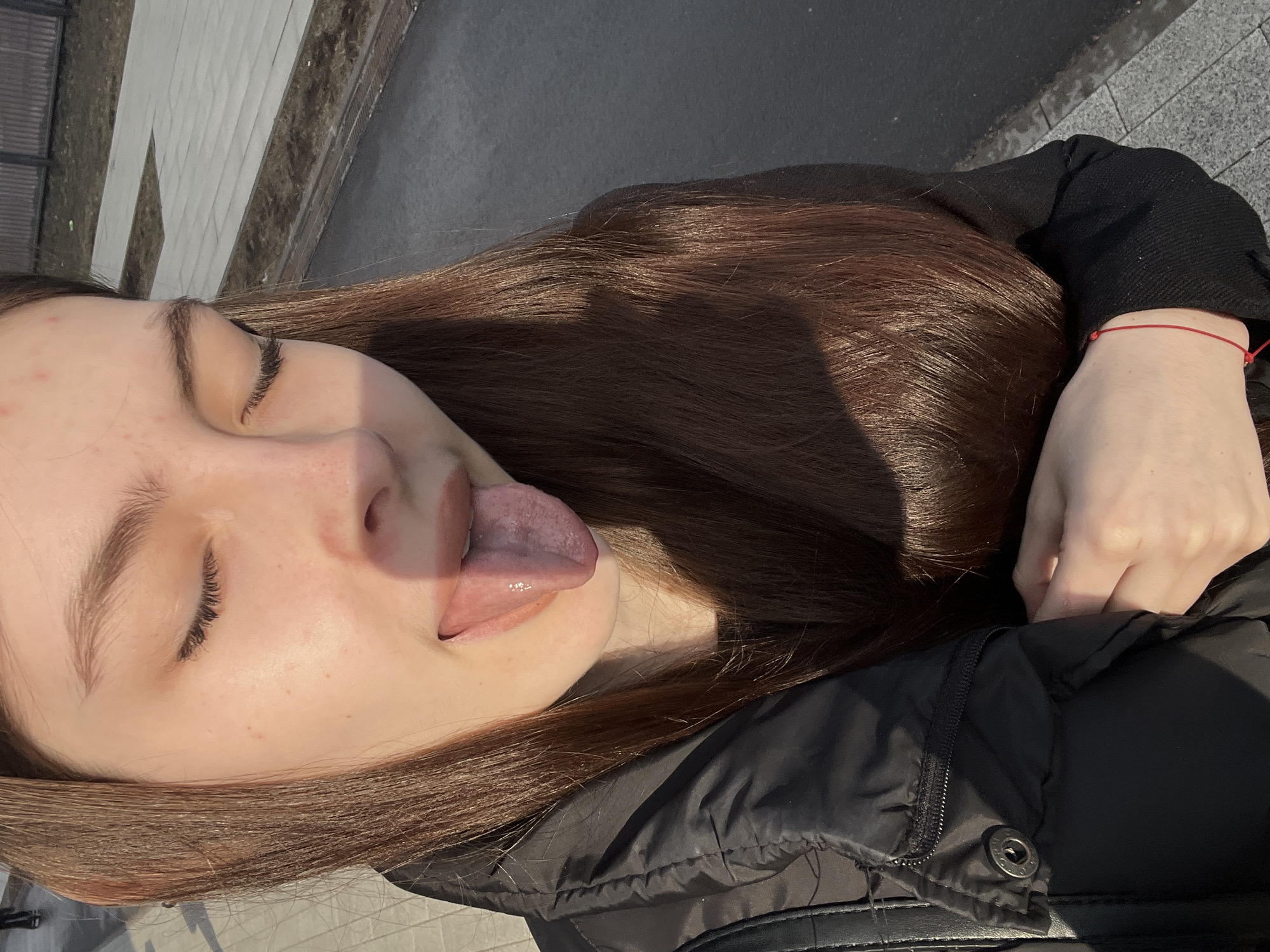 A young woman with brown hair is visible in the frame. She is wearing a black jacket and a red string bracelet on her left wrist. Her eyes are closed, and her tongue is extended. The scene appears to be outdoors, with a sidewalk and a building visible in the background. The location may be the city of Saint Petersburg, Russia.