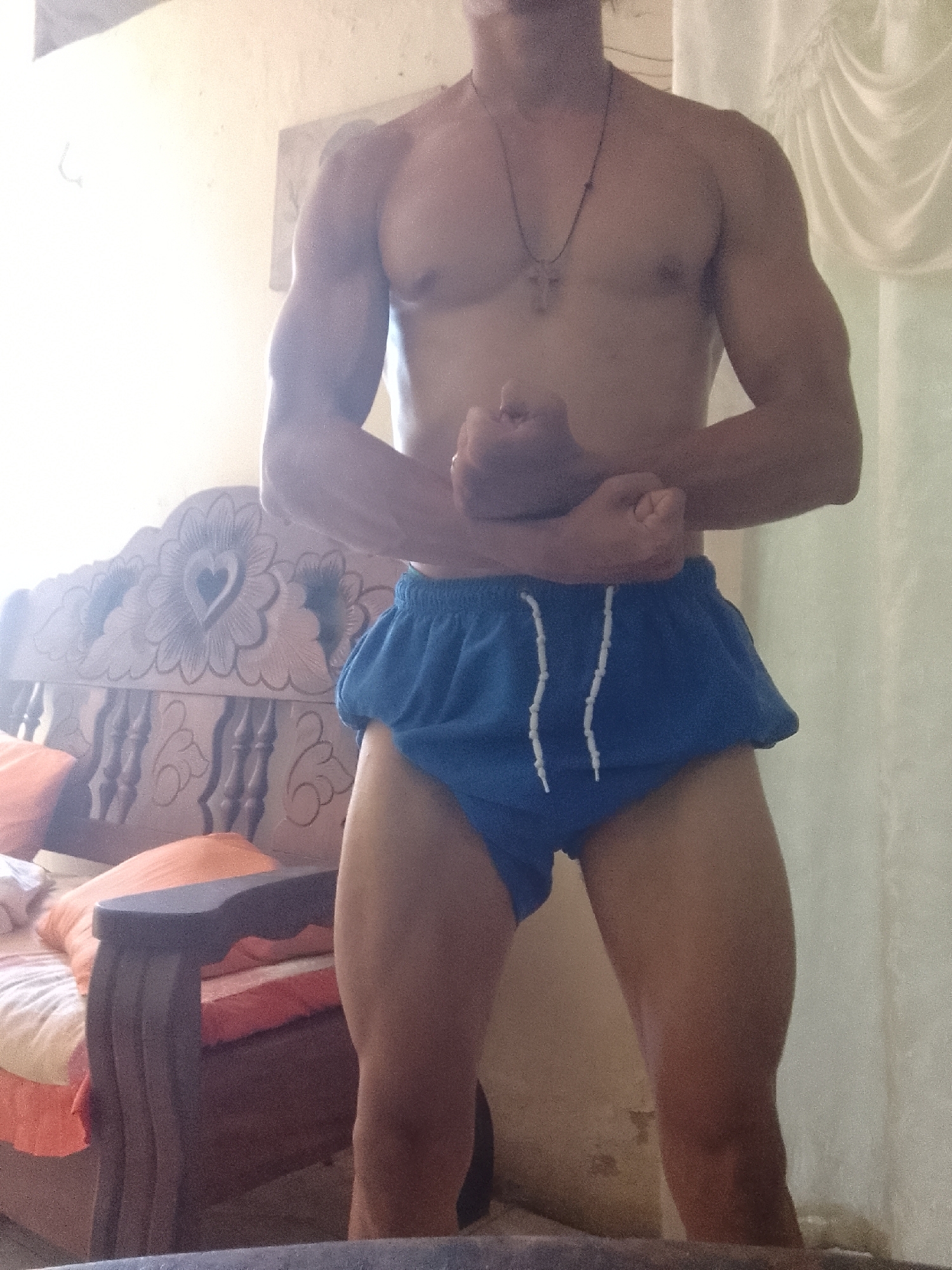 A medium shot depicts a shirtless man with a muscular build, wearing blue shorts with a white drawstring. He is flexing both arms. The man wears a necklace with a cross pendant. Behind him is a wooden bed frame with a heart carving on the headboard. Orange pillows are visible on the bed. A sheer white curtain hangs on the right side of the frame. The background wall is light colored. Based on the style of the room's furniture and interior decor, this image could plausibly have been taken in a residential setting in a city like Juan Griego, Venezuela, however, without further information, the specific location can't be determined for certain.