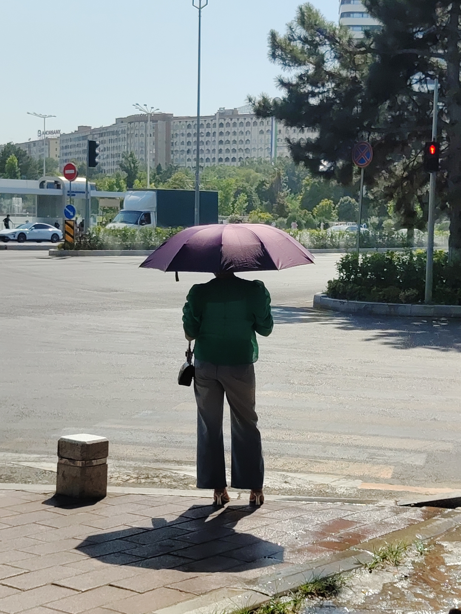 In the city of Tashkent, Uzbekistan, on a bright, sunny day, a person stands on a wet crosswalk holding a closed, dark purple umbrella over their head, shielding them from the sun. The individual is wearing a green jacket, gray pants, and heeled shoes. They are carrying a black purse. In the background, a white car and a white truck are visible on the road. Several buildings, including one with the prominent sign "AMORBANK," line the horizon. A traffic light shows red, and other traffic signs are present. The scene suggests the pavement may have been recently wet, perhaps from sprinklers or a brief shower, as evidenced by the reflections and pooling water.
