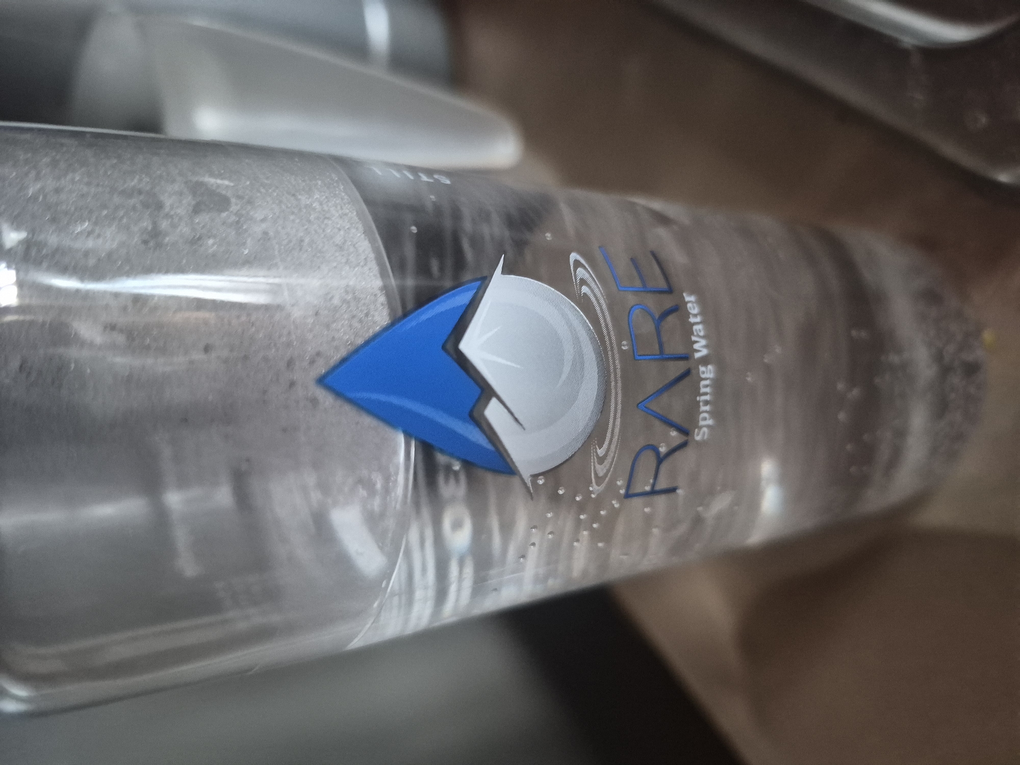 The image displays a clear plastic water bottle, angled diagonally, filled with transparent liquid. Numerous small bubbles are visible clinging to the inside surface of the bottle. The bottle's label features a distinct logo: a blue, teardrop-shaped element pointing left, alongside a white, circular element containing an arrow pointing right. The brand name "RARE" is prominently printed in dark blue, stylized capital letters, with "Spring Water" written in smaller text below it. Faintly visible through the bottle, likely on the reverse side of the label, is the word "STILL". The background is blurred, indicating an indoor setting, possibly a counter or table. The scene depicts a still object; there are no human subjects or activities taking place, and no specific time of day or weather cues are present. The image itself does not contain visual markers for its location in Yerevan, Armenia.