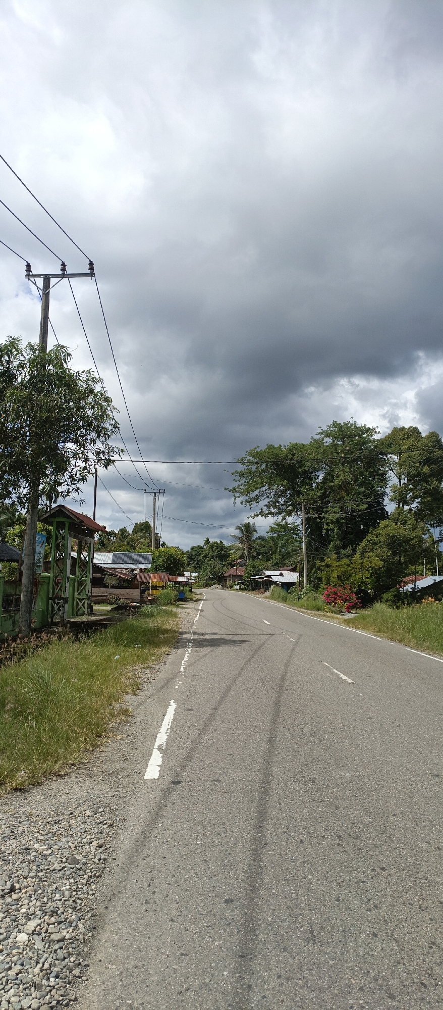 This image depicts a paved road in a rural or semi-rural area, likely Iemirah, Indonesia. The sky is overcast with dark, heavy clouds. On the left side of the road, there is a gravel shoulder with tall grass. A wooden utility pole with electrical wires runs along the side of the road. Adjacent to the pole is a green, open-sided structure with a rusted roof, possibly a bus stop or shelter, with some signage. Further down the road on the left, several small buildings with corrugated metal roofs are visible. The road itself has tire marks and a broken white line marking the lane. On the right side of the road, there are more trees and vegetation, with several houses visible in the distance. The overall scene suggests a cloudy day in a developing area with basic infrastructure. There are no people or animals visible in the image.