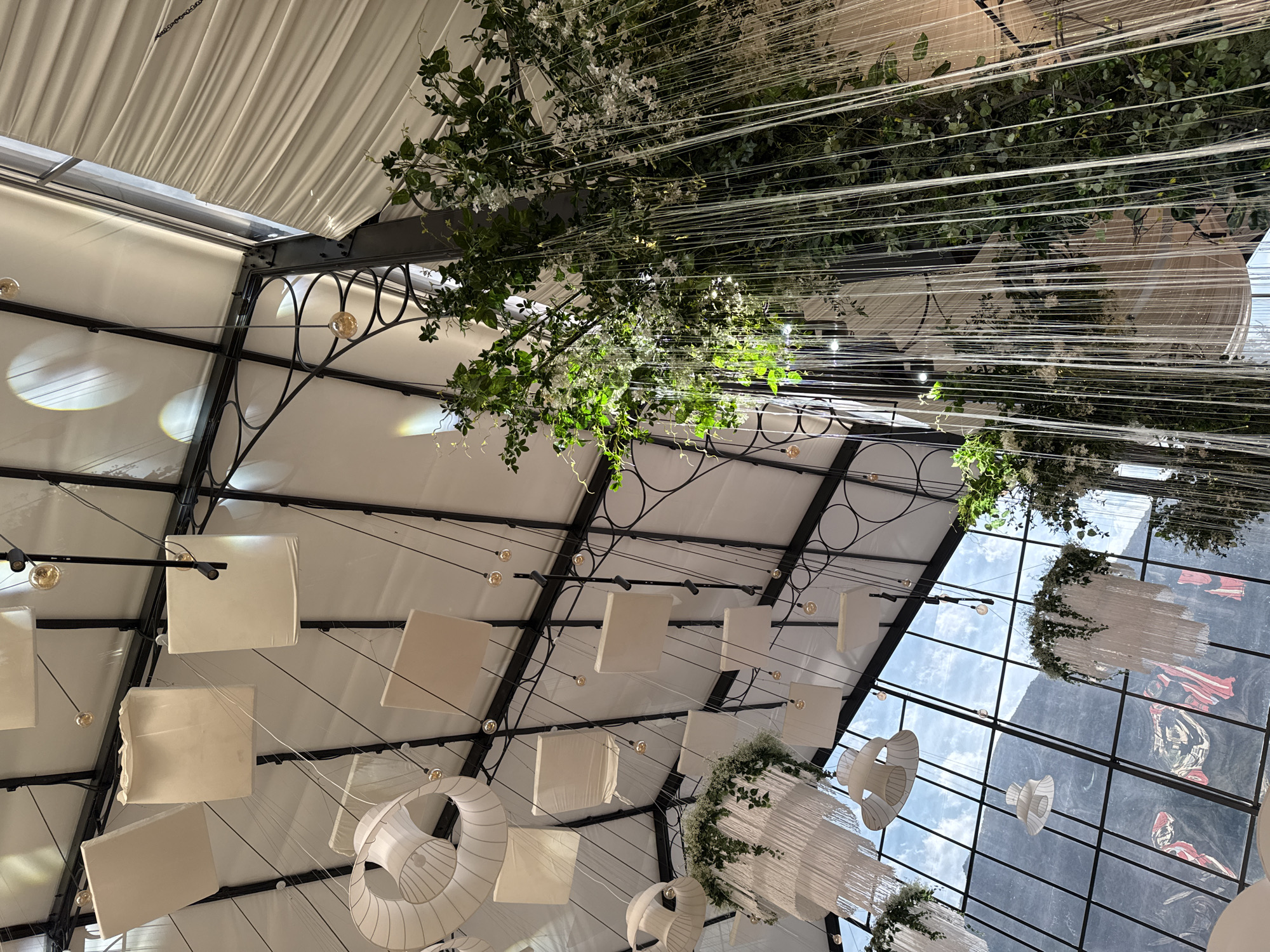 The image depicts an indoor space with a high, vaulted ceiling constructed of white fabric and metal framework. The ceiling is adorned with numerous hanging elements, including square white panels, spherical glass light fixtures, and elaborate white chandeliers decorated with greenery and fringe. A prominent feature is a large, vertical installation of dense green foliage with cascading white flowers and delicate, translucent strands, suggesting a lush, naturalistic aesthetic. Below the ceiling, large glass windows reveal a backdrop of mountains under a partly cloudy sky. The architectural style appears to be modern, possibly a venue or event space, designed to evoke a sense of ethereal beauty with its combination of natural and decorative elements. The location is identified as Arashan, Kyrgyzstan, indicating a specific setting within a mountainous region.