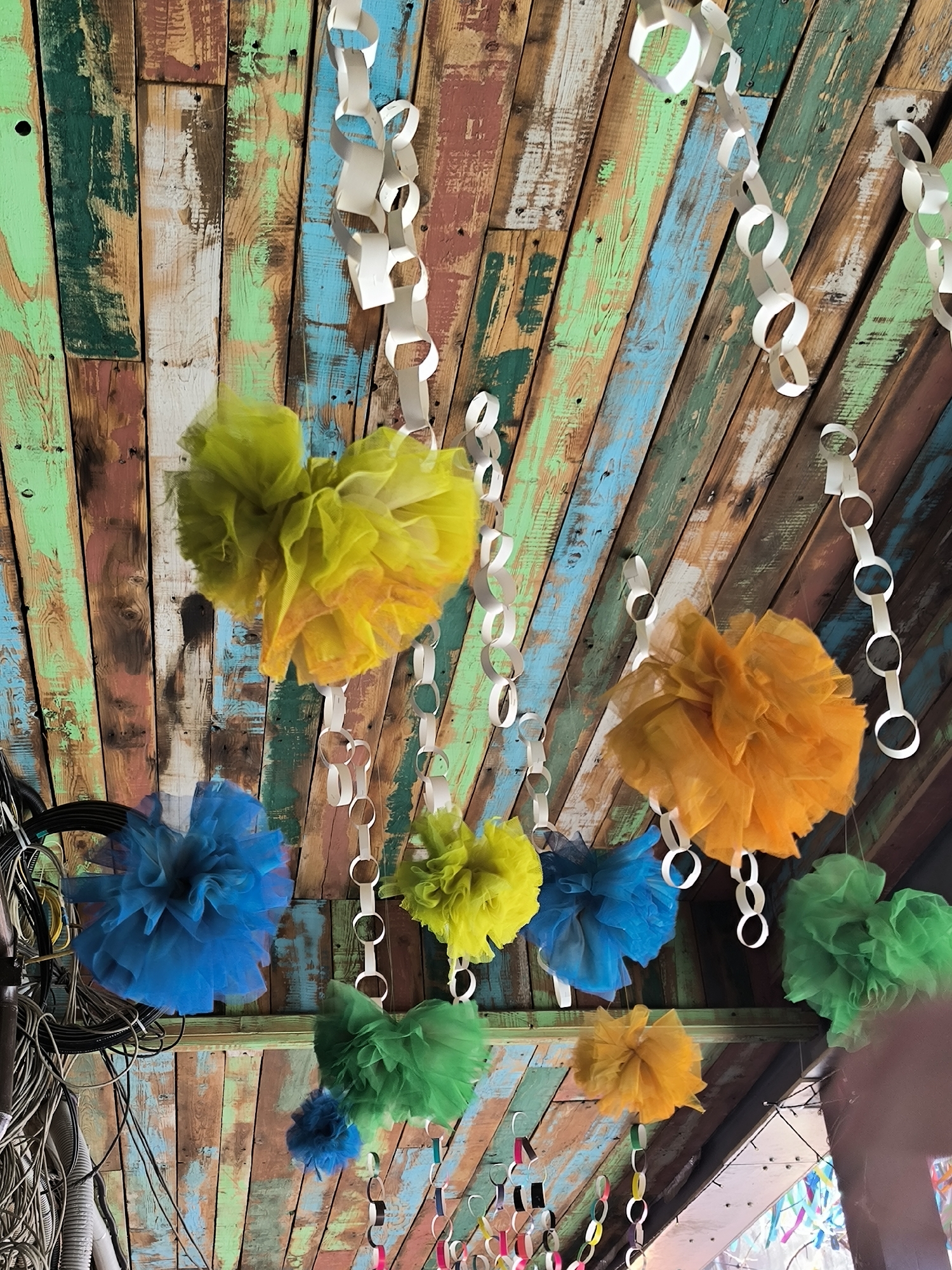 The image shows a colorful ceiling made of reclaimed wood planks painted in various shades of green, blue, brown, and white. Several decorative items are hanging from this ceiling. These include white paper chains and numerous fluffy pom-poms in bright colors such as yellow, orange, blue, and green. Some of the pom-poms appear to be made of tulle or a similar sheer fabric, giving them a light and airy appearance. In the lower left corner, there is a bundle of black electrical wires and cables, which contrasts with the festive decorations. The overall impression is one of a celebratory or decorative space, possibly an outdoor area or a casual indoor venue. The lighting suggests it could be daytime. There is no visible text in the image.