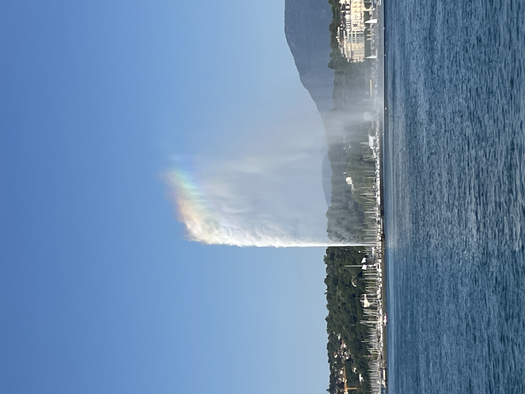 This image captures the Jet d'Eau, a famous landmark in Geneva, Switzerland, erupting water high into the clear blue sky. A faint rainbow is visible within the spray of the fountain, refracting the sunlight. The Jet d'Eau is situated in the middle of Lake Geneva, with the city's shoreline visible in the background. The shoreline is lined with a dense green forest, dotted with buildings that appear to be houses and hotels. A large number of sailboats are moored along the waterfront, their masts creating a distinctive silhouette against the land. The water of the lake is a deep blue, with small ripples on its surface. The overall scene depicts a sunny day with a gentle breeze, creating a picturesque view of the lake and its iconic fountain.