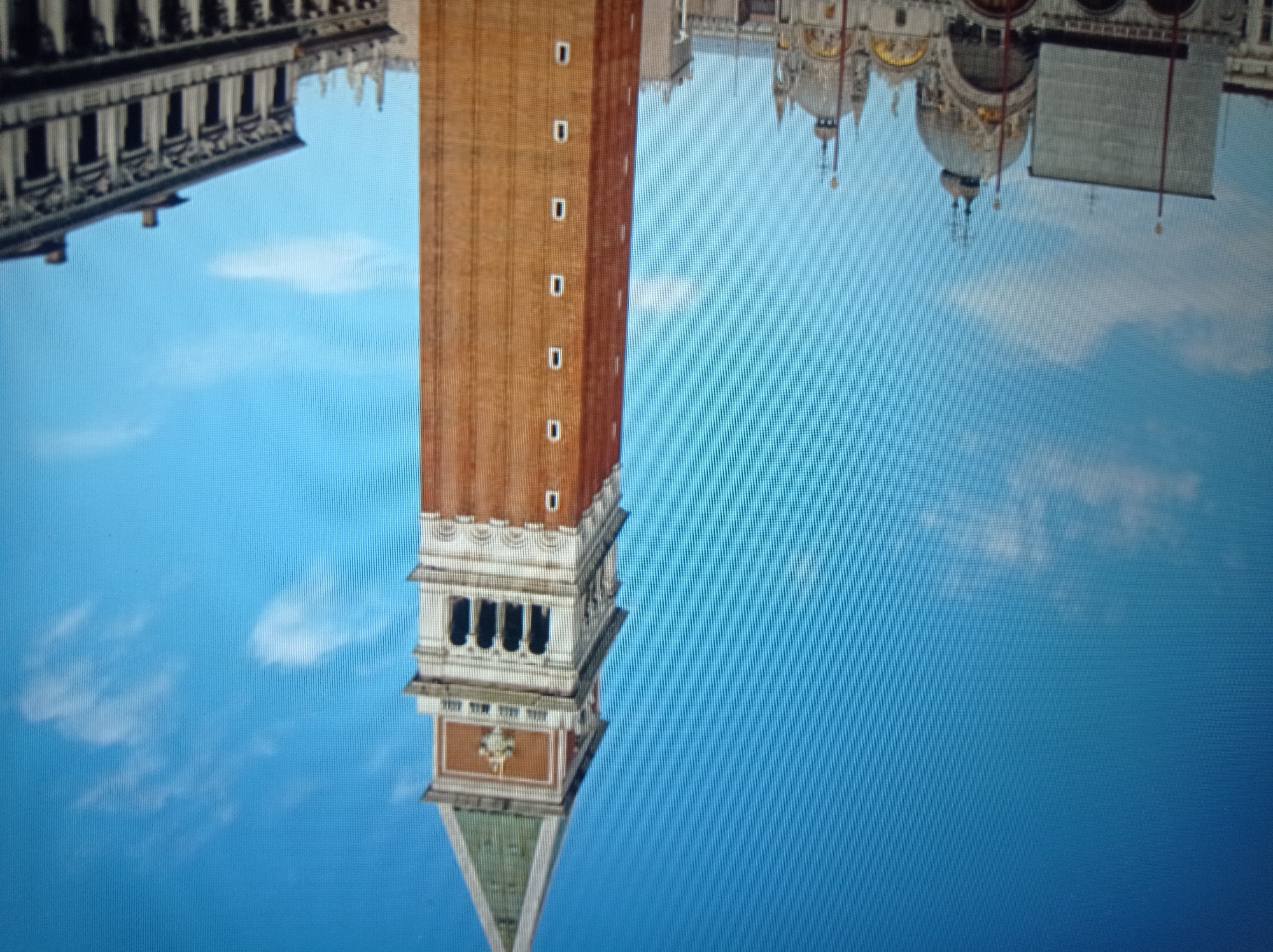 The image shows a tall brick tower with a green pyramidal roof against a blue sky with scattered white clouds. The tower has several rectangular windows and ornate architectural details near its top. To the left of the tower, a large building with domes and intricate facade is visible. To the right, another building with a classical architectural style, featuring columns and multiple windows, is present. The scene appears to be an outdoor view of historical buildings in a city. The provided location is Mukachevo, Ukraine.