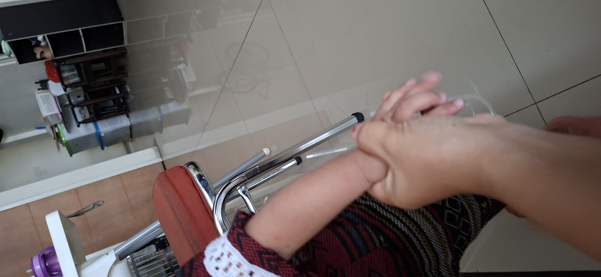 The image depicts a close-up view of a person holding a child's arm. The child is wearing a red and black patterned garment with white trim on the sleeve. The adult's hand is gently grasping the child's forearm. In the background, a red-seated chair with chrome legs is visible. Further back, a white high chair with a purple sippy cup is present. The floor is tiled in a light neutral color, reflecting the surroundings. The environment appears to be an indoor domestic setting, possibly a living room or dining area. The image was taken in Surabaya, Indonesia.