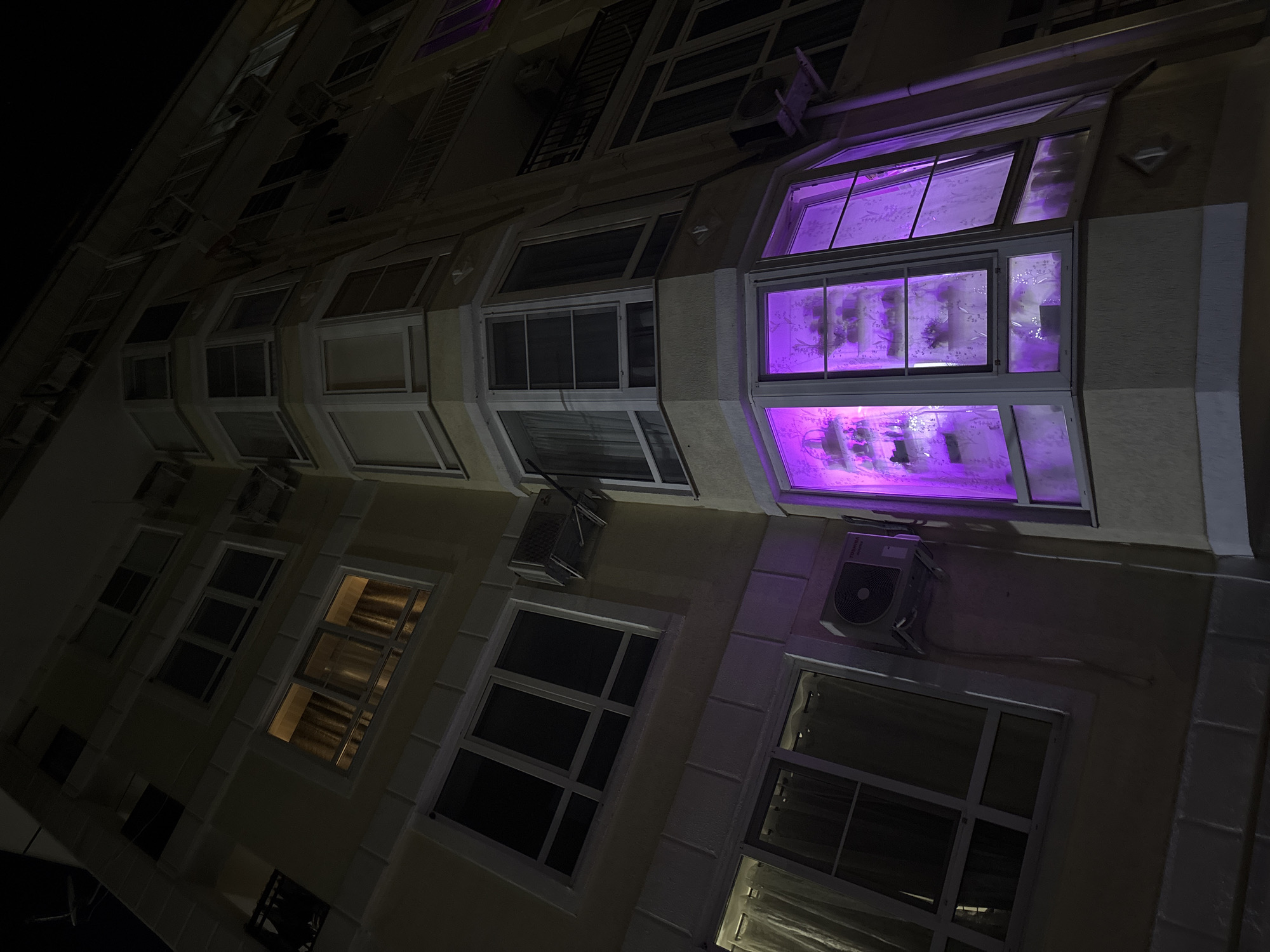 The image depicts the exterior of a building at night. The architecture is modern, with multiple windows and air conditioning units. One window is illuminated with a bright purple light. The building appears to be located in an urban area, likely the city of Sochi, Russia. The photograph is taken from a low angle, looking upwards. No people are visible.