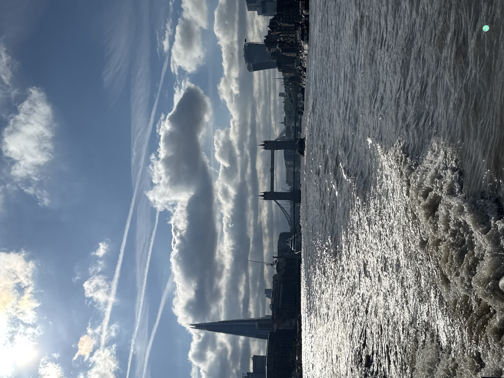 The image shows a view of the River Thames in London, United Kingdom, on a cloudy day. In the background, the iconic Tower Bridge is visible, along with The Shard and other modern skyscrapers. The sun is shining through the clouds, creating a bright reflection on the water. A boat is leaving a wake as it moves through the choppy river.
