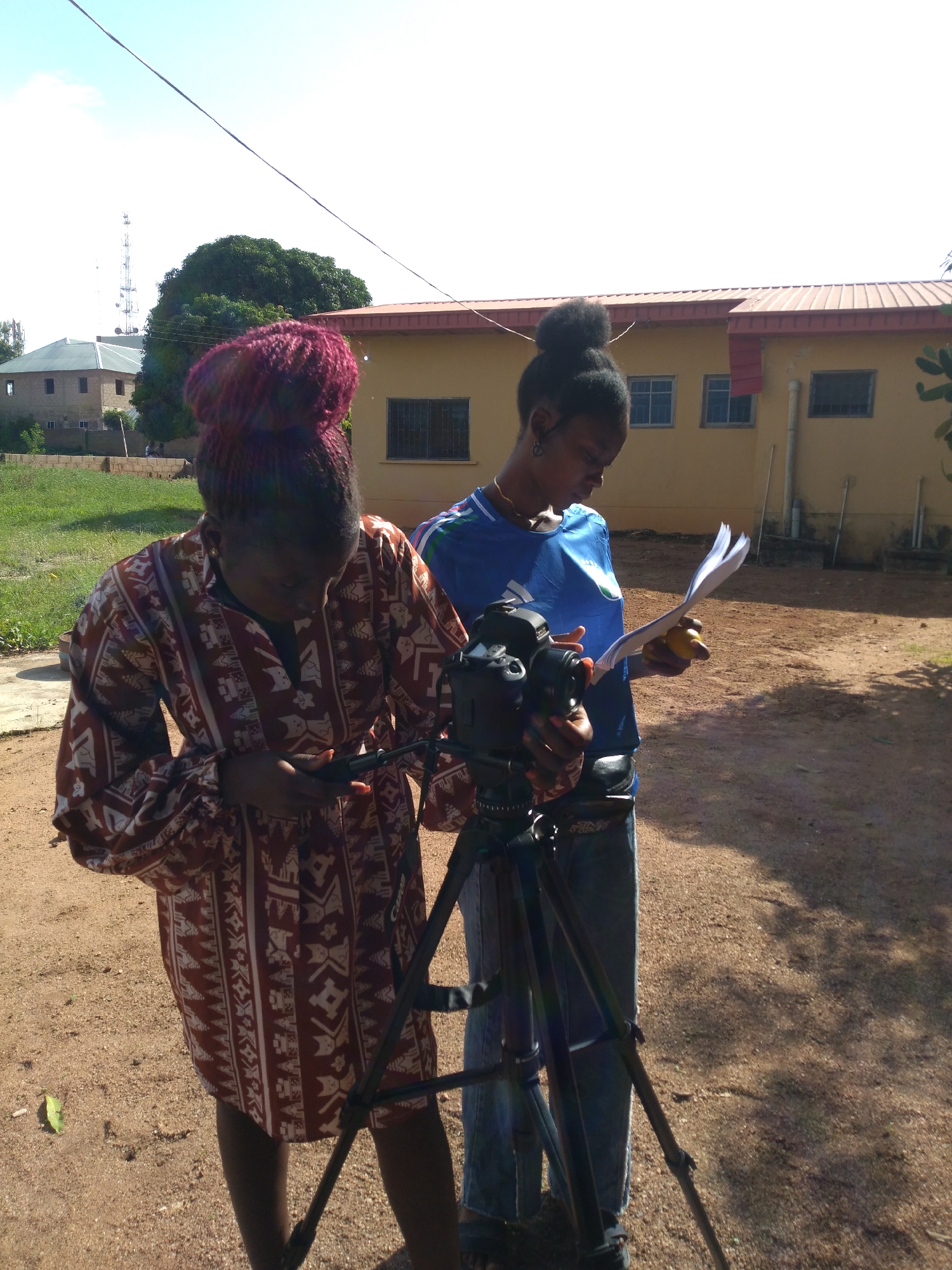 This image captures an outdoor scene in Jos, Nigeria, under a clear, bright sky. Two individuals are central to the frame, standing on reddish-brown dirt ground.
The person on the left has dark skin and burgundy dreadlocks or braids styled into a high bun. She wears a long-sleeved, patterned tunic-style top or dress in reddish-brown and white, featuring geometric and possibly abstract animal motifs. Her gaze is directed downwards as she holds a small, black rectangular electronic device in both hands.
The person on the right, also with dark skin, has her dark hair pulled into a high bun. She is dressed in a blue t-shirt with a white logo (possibly Adidas) and light blue jeans. She wears a thin gold-colored necklace and small hoop earrings. She is operating a black DSLR camera, which is mounted on a black tripod, with her left hand. In her right hand, she holds and reviews a stack of white papers. Both individuals are focused on their respective tasks, appearing to be involved in a collaborative filming or photography setup.
The background features a single-story, light yellow building with a reddish corrugated metal roof on the right. On the left, a two-story, light-colored building with a grey sloped roof is visible. A large, dense green tree stands between these buildings. In the far distance on the left, several tall metal antenna or communication towers rise against the clear blue sky. A dark, thin cable or power line stretches diagonally across the upper portion of the image. The ground is loose, dry dirt with scattered small stones, and a single green leaf rests on the ground in the lower-left.