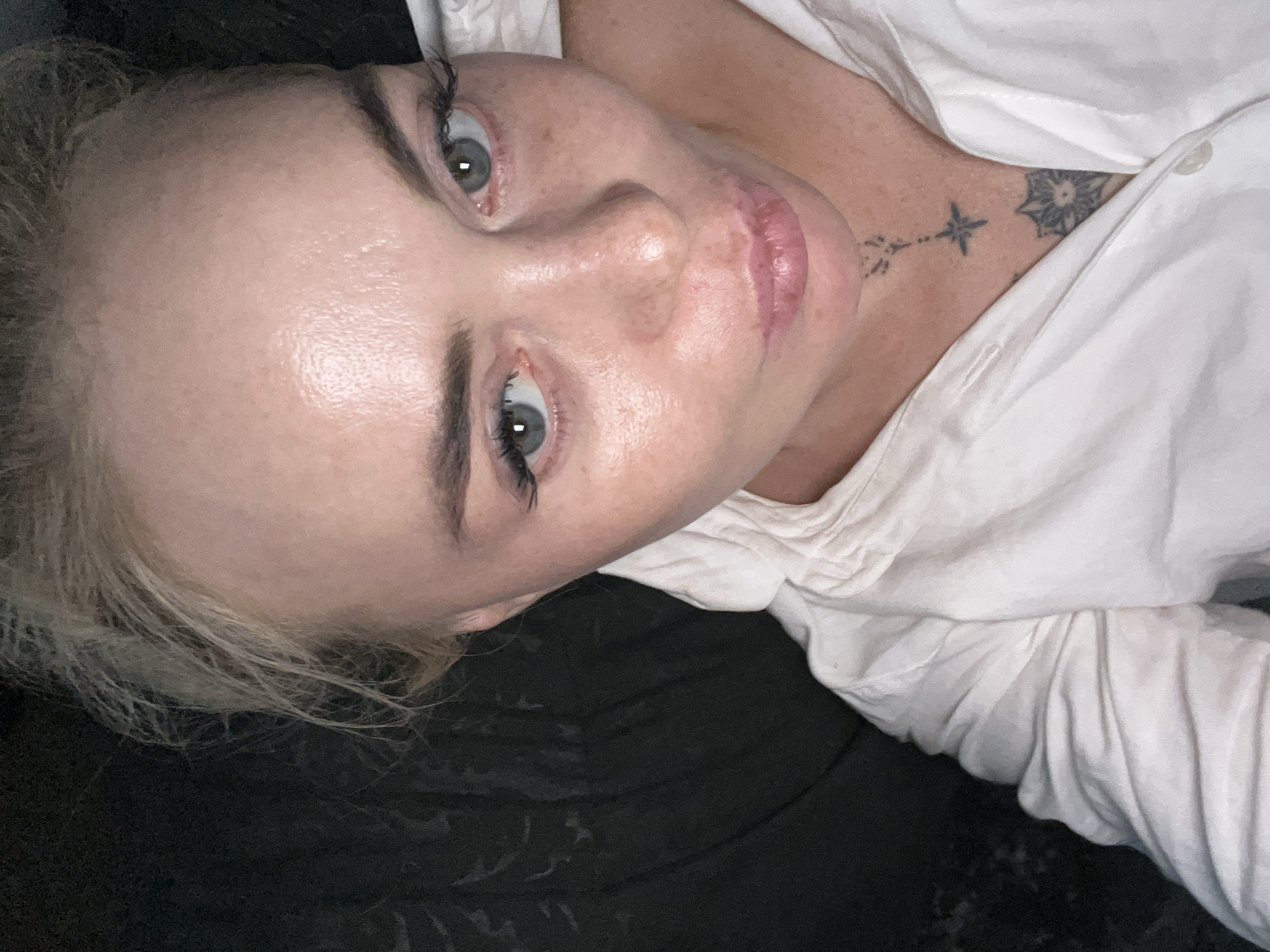A close-up, high-angle selfie shows a fair-skinned person with blonde hair pulled back, wearing a white collared shirt. The subject's face is illuminated by a light source, highlighting their blue-grey eyes, full lips, and fair skin with freckles. The background is dark and indistinct, with the suggestion of a textured surface on the left side. A small, dark tattoo is visible on the person's neck, just above the collarbone. The overall impression is an intimate, casual portrait. The image does not provide any clues about its specific location in the city of Unknown, Unknown.