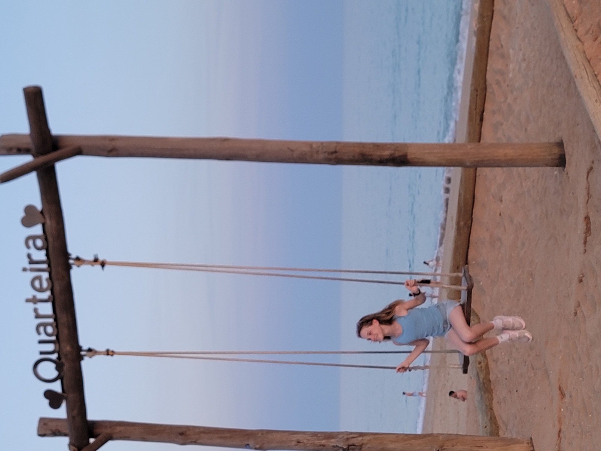 A young girl is on a swing at the beach in Quarteira, Portugal. She is wearing a blue tank top and light-colored shorts with white sneakers. Her hair is long and brown, and she is smiling as she swings. The swing set is made of wood and has a sign that reads "Quarteira" with a heart shape. The beach is sandy, and the ocean is visible in the background. The sky is a soft blue, suggesting it is either early morning or late evening. Other people can be seen in the background on the beach. The overall mood of the image is peaceful and joyful.