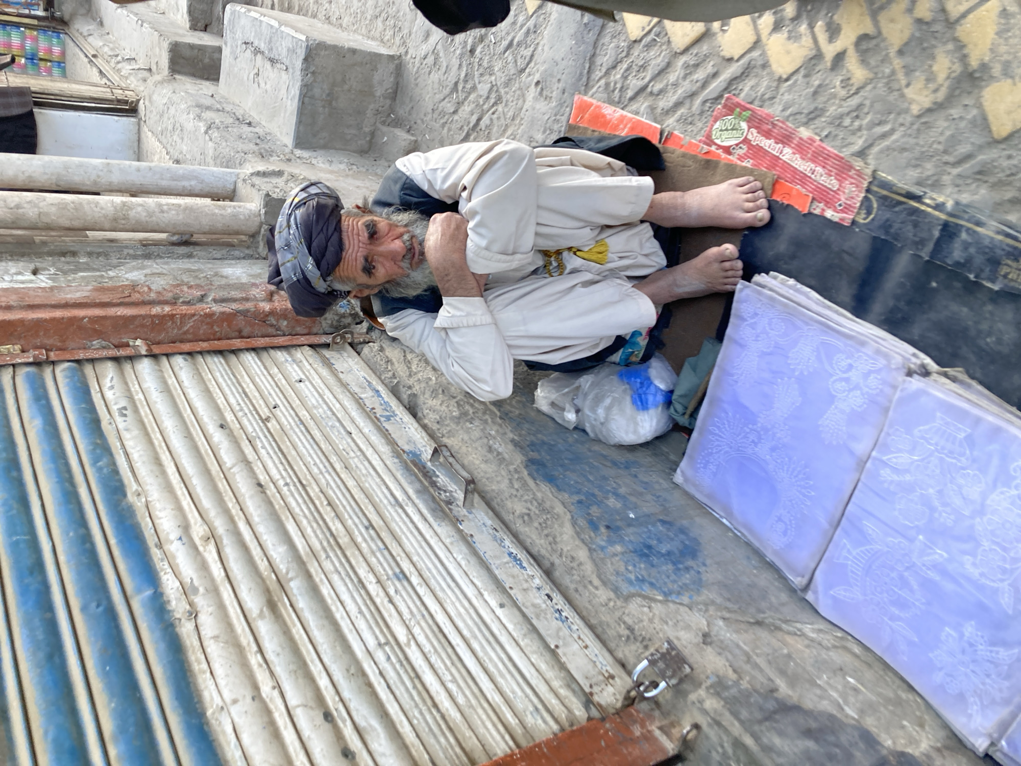 The image shows an older man lying on what appears to be a corrugated metal surface, possibly a shop shutter, in Quetta City Tehsil, Pakistan. He is wearing traditional clothing, including a turban, and has a weathered appearance. He is partially covered by cardboard. Next to him are stacks of what appear to be fabric or embroidered items wrapped in plastic. The surroundings suggest a cluttered urban environment, with concrete steps and other objects in the background.