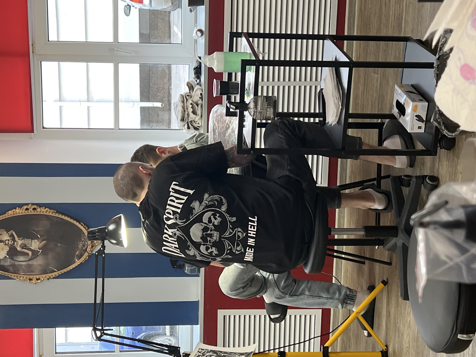 The image shows two men in what appears to be a tattoo studio in Saint Petersburg, Russia, during daytime. The scene is brightly lit, primarily by natural light streaming through a large window.
In the foreground, a man sits on a black wheeled stool, facing away from the viewer. He is wearing a black t-shirt featuring a prominent white graphic design on the back that includes the text "DARK SPIRIT" across the top, a large spade symbol with multiple skulls inside, and "MADE IN HELL" written vertically on the right. He wears light blue jeans and black slides with white socks. A yellow stand, likely part of a task lamp, is visible near his feet.
Another man is partially visible opposite him, suggesting he is either a client or another tattoo artist. Between the two men is a black metal rolling cart used for tattoo supplies. On the cart, there is a green liquid in a squeeze bottle, a clear glass container, a black cup, and white paper towels. A white box labeled "GLOVES" is placed on a lower shelf of the cart.
A bright black task lamp with a large, adjustable head is positioned to illuminate the workspace. The wall to the left features horizontal blue and white stripes, with an ornate oval gold-framed picture depicting a woman hanging on it. A plain red accent wall is visible on the right. Through a white-framed window, an exterior view of what appears to be a street or courtyard is visible under bright daylight. The floor is covered with light-colored wooden or laminate planks. The arrangement of equipment and individuals strongly suggests that a tattoo session is either underway or about to commence.