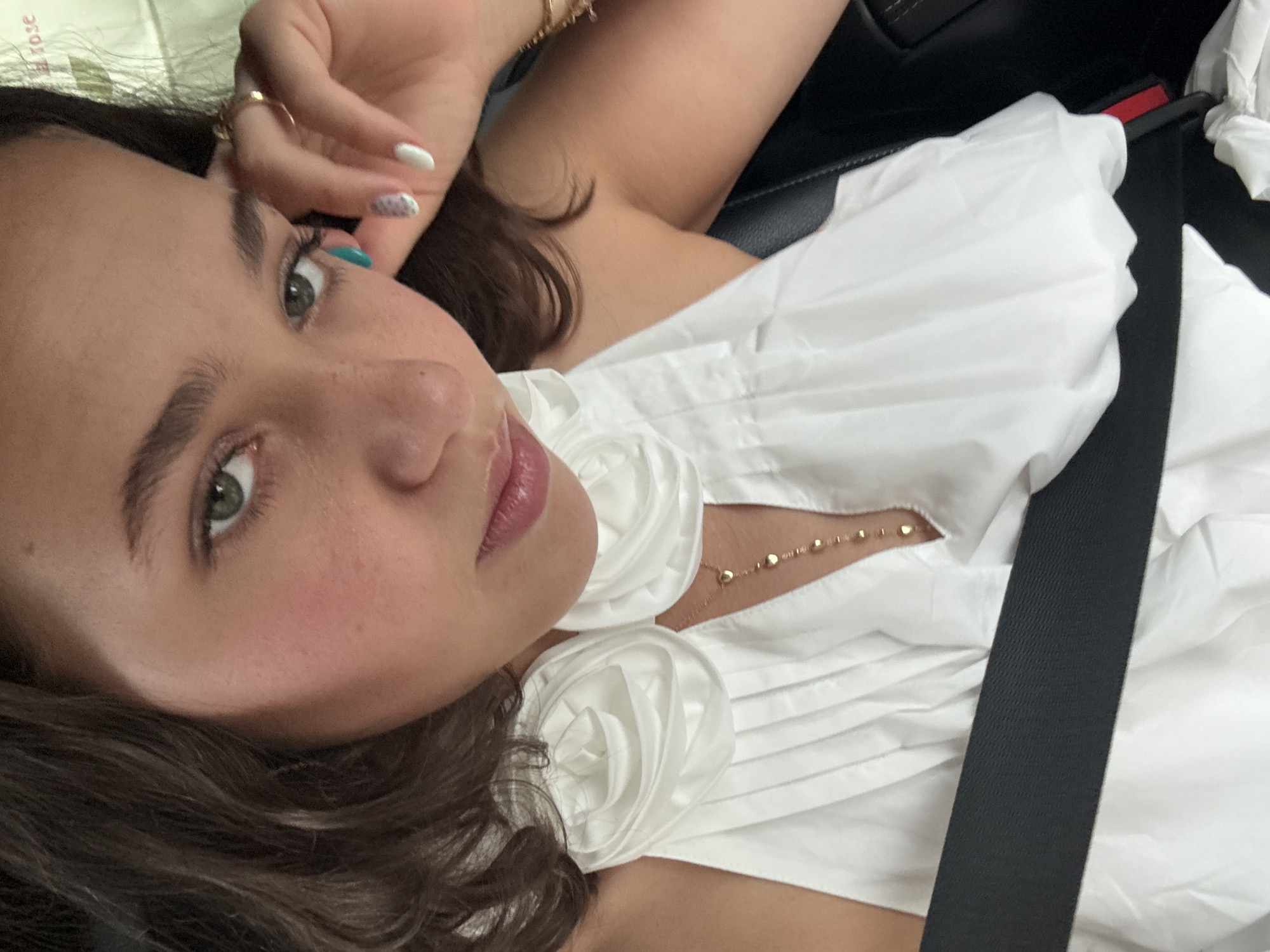 A medium shot shows a young woman with dark wavy hair and green eyes, wearing a white dress with fabric roses on the shoulders, seated in a car, possibly in Milan, Italy. She has light makeup, with a hint of blush on her cheeks, and her lips are slightly parted. Her right hand is raised to her face, touching her temple, and she is wearing rings and a bracelet. Her fingernails are painted with a white and teal design. A gold necklace with small heart-shaped pendants is visible against her dress. A black seatbelt crosses her chest. The background is blurred, but hints of a headrest and a subtle rose-colored text can be seen, possibly indicating a car interior or personal item.