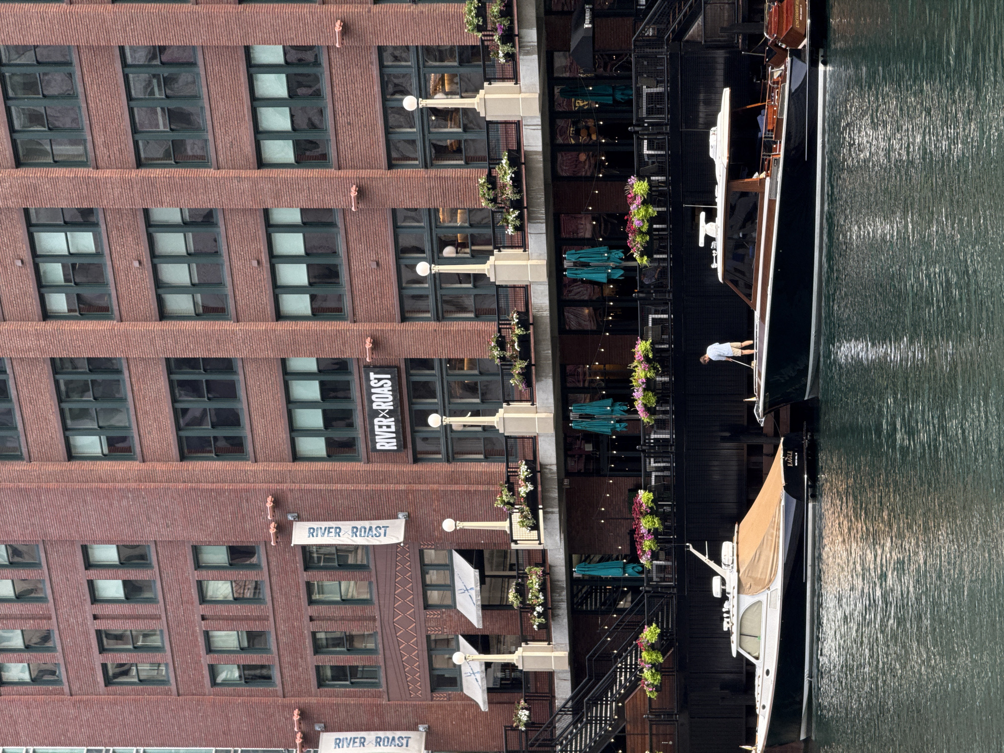 The image depicts a waterfront scene in Chicago, Illinois, USA, likely along the Chicago River. In the foreground, two elegant motor yachts are docked. The yacht on the left, named "EAGLE," is dark-hulled with a tan cover over its forward section. The yacht on the right is dark-hulled with prominent wooden trim. A lone individual in a light-colored shirt and shorts is visible on the deck of the right yacht, appearing to be cleaning or attending to it with a long-handled tool.
Behind the yachts, a multi-story brick building rises, featuring numerous dark-framed windows. The building's facade is adorned with white banners displaying the "RIVER ROAST" logo. A raised walkway or balcony runs along the second story of the building, decorated with planters filled with vibrant flowers. This walkway overlooks a lower level with outdoor seating, awnings, and umbrellas, suggesting a restaurant or establishment. The water of the river is calm, reflecting the overcast sky and the surrounding architecture.