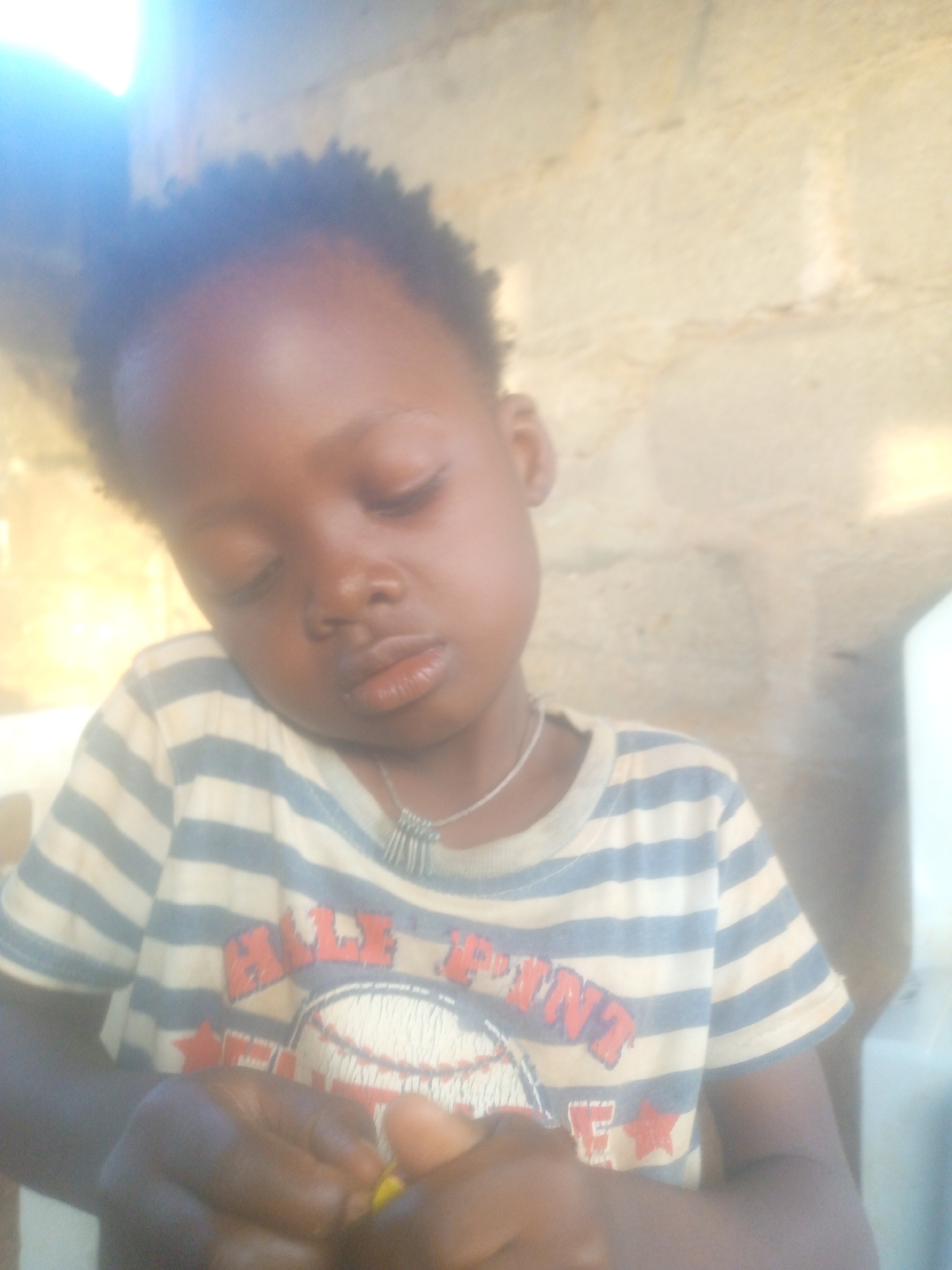 A young Black child with short, curly hair is sitting indoors, eyes closed or downcast. They are wearing a striped t-shirt with a baseball graphic and a silver necklace. Their hands are clasped together in front of them, possibly holding a small yellow object. The background is a light-colored wall. The location is Unknown, Unknown.