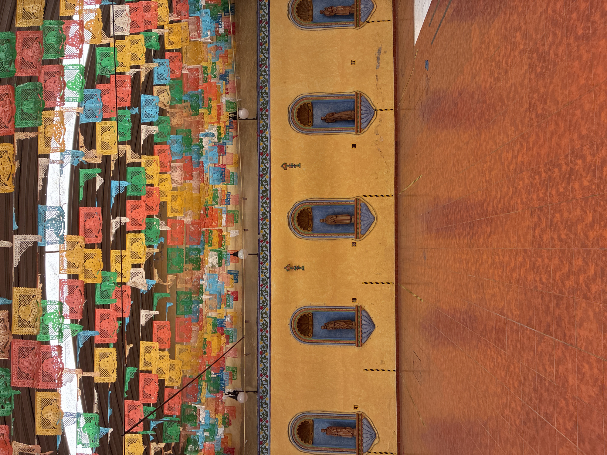 The image shows an upward, angled view of a building facade in Puebla, Mexico. The left side is decorated with rows of colorful paper cutouts (papel picado) suspended from the ceiling, casting dappled light. Adjacent to the paper decorations is a tan-colored wall with several alcoves containing statues. Numbers are painted near the alcoves. The wall is adorned with floral detailing. To the right is a tiled reddish-brown wall.