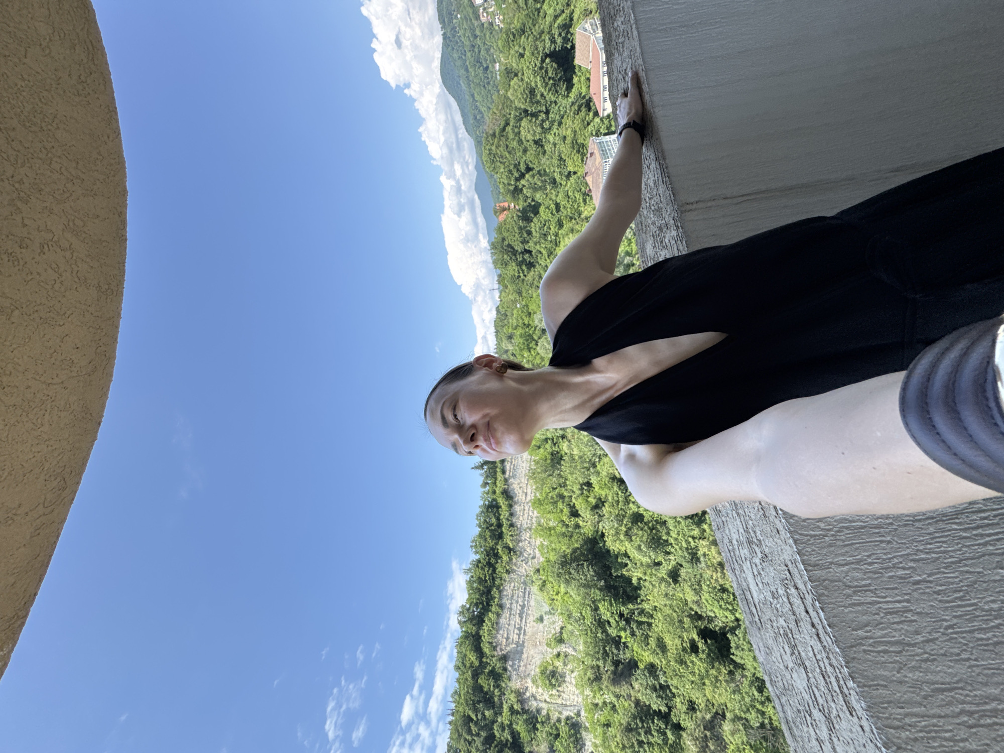 A young woman with her hair pulled back is taking a selfie on a balcony. She is wearing a black V-neck dress and is leaning against the railing, looking up at the sky with her eyes closed. The background shows a lush green mountainous landscape with some buildings visible in the distance under a bright blue sky with scattered clouds. This scene is consistent with a location in Sochi, Russia, known for its mountainous terrain and proximity to the Black Sea.