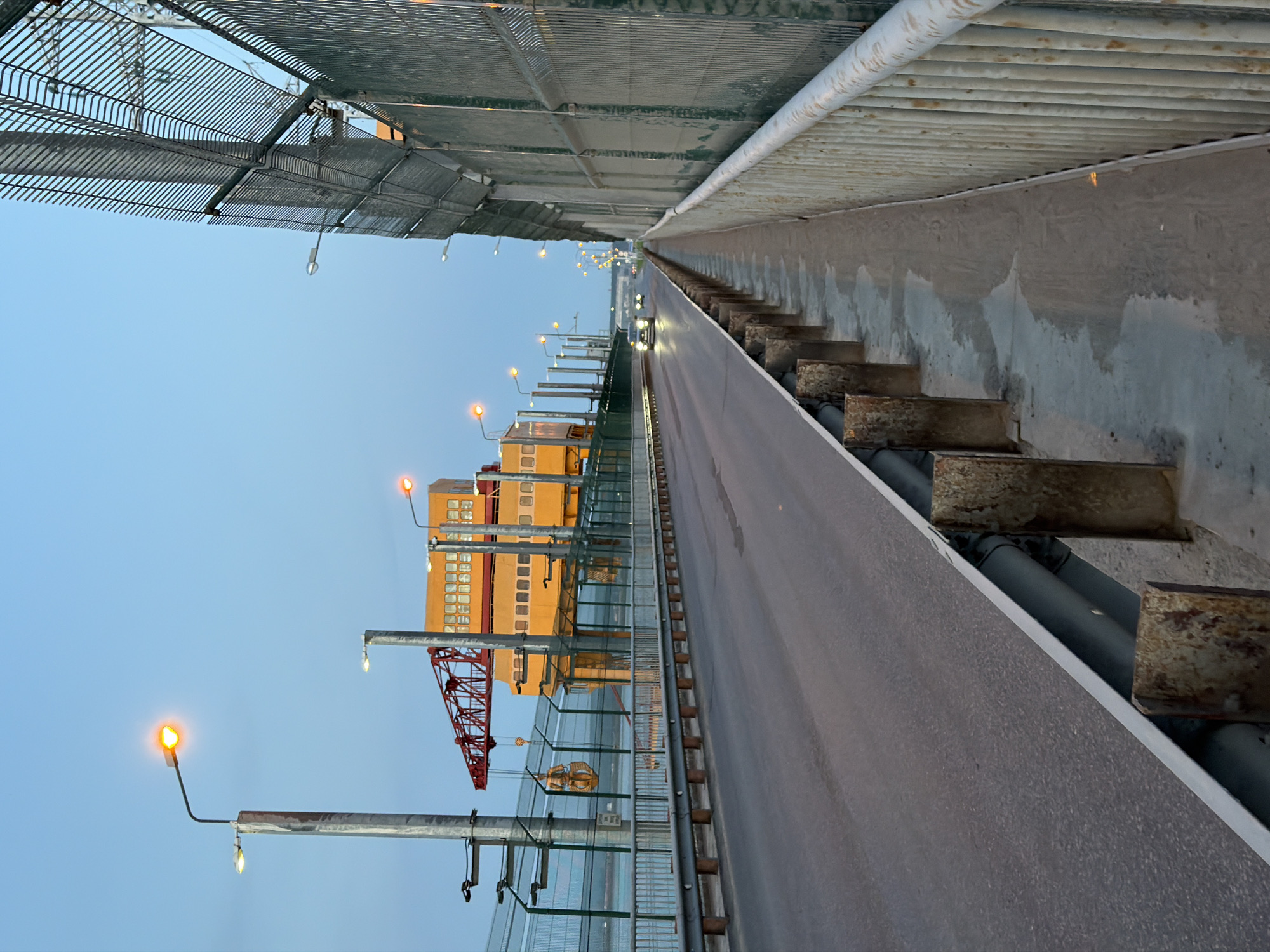 This image depicts a road or bridge in the city of Chaikovsky, Russia. The viewpoint is from the side of the road/bridge looking lengthwise. On one side of the road/bridge there's a fence and lampposts with orange lights leading to a tall yellow building and a construction crane in the background. The other side of the road/bridge features concrete steps leading up to a higher level, with grey pipes running parallel to the road/bridge. Above, a wire mesh ceiling structure is visible. In the distance, two vehicles with headlights on appear on the road. The sky above is clear and light blue.