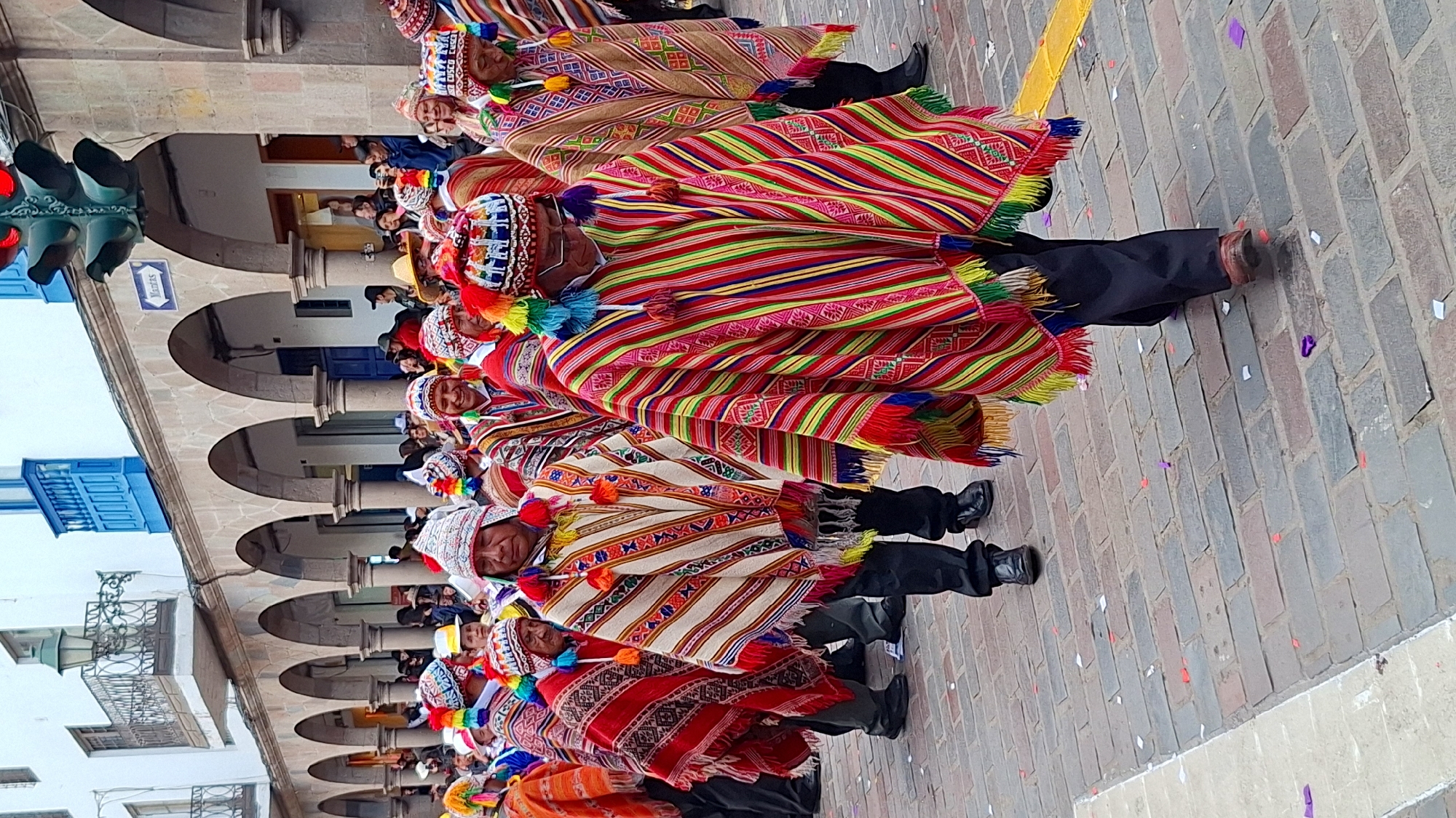 This image captures a vibrant cultural procession or parade taking place outdoors in a public square in Cuzco, Peru. A long line of men, dressed in traditional Andean attire, moves across a cobblestone pavement. Each man wears a strikingly colorful poncho featuring bold stripes and intricate geometric patterns in shades of red, green, yellow, blue, and white, adorned with tassels around the shoulders and hem. They also wear distinctive white hats or headwear with colorful patterns and pompoms. Black trousers and dark shoes complete their outfits. Many of the participants are smiling, contributing to a festive atmosphere.
The ground is paved with varied shades of grey and reddish-brown bricks, scattered with small pieces of colorful confetti (purple, white, red, yellow), indicating a celebration. A yellow line is visible on the pavement. In the background, to the left, are colonial-era buildings with white facades and repetitive arched doorways and windows, suggesting a historic city center. Some onlookers can be seen under these arches. A blue traffic light with a red light illuminated is visible on the far left. Above the arches, a blue rectangular sign is partially visible, with the Spanish word "Ventanías" and an arrow. The scene is brightly lit, suggesting a clear daytime event under sunny weather.