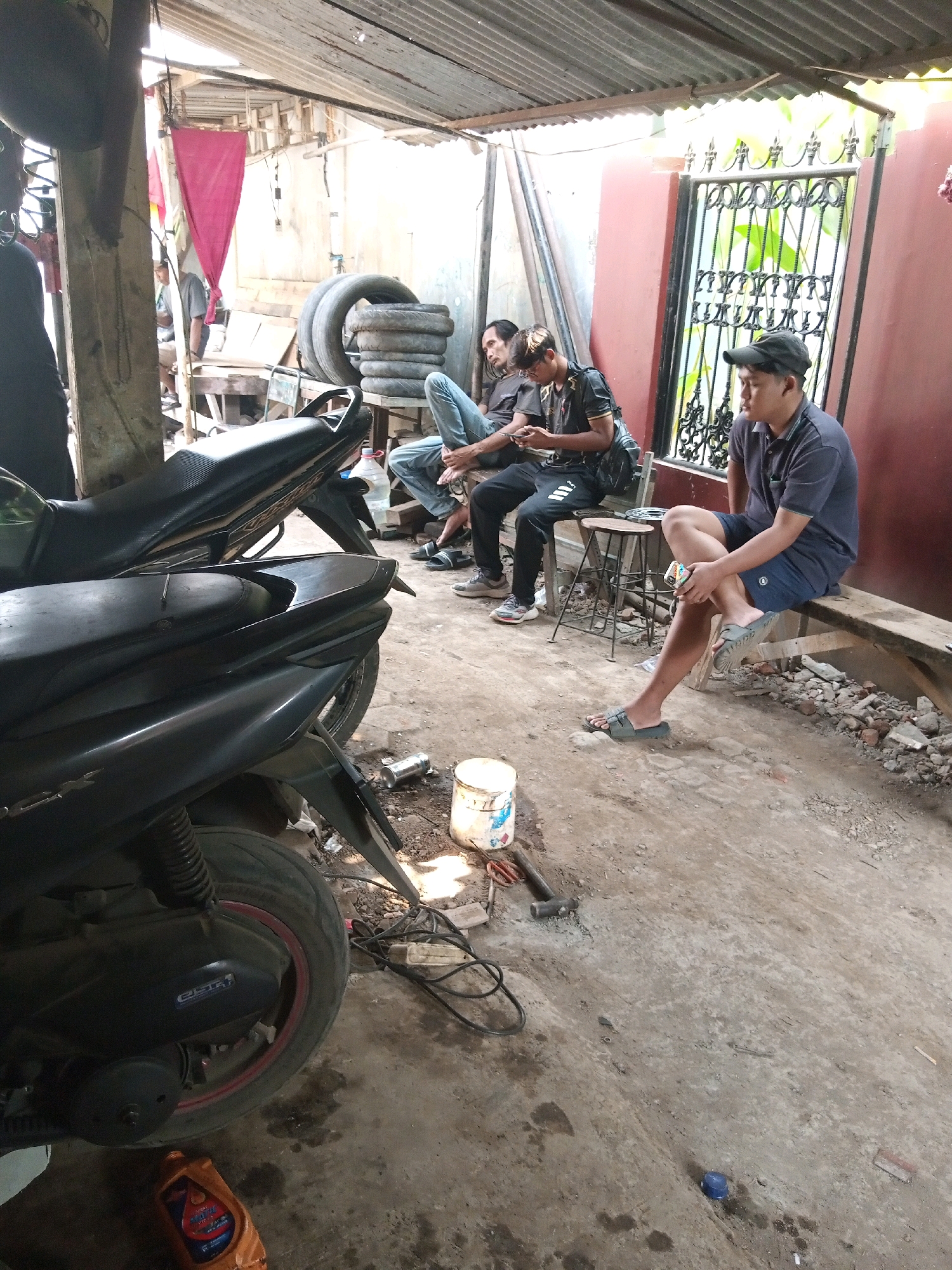 In Batang, Indonesia, a group of men are gathered in an outdoor, semi-covered workshop. Two motorcycles, a scooter and a sportbike, are parked in the foreground. The scooter is black and has a red rim on its rear wheel. The sportbike is also black.
The environment appears to be a repair shop, with stacks of tires visible in the background, along with a red curtain and corrugated metal roofing. Tools like a hammer and a wrench are scattered on the dirt floor, along with a white bucket and a bottle of orange oil.
Several men are seated or standing around. One man in a blue polo shirt and dark shorts is sitting on a wooden bench to the right, holding a cigarette. Another man, wearing a dark t-shirt and black pants, is seated on a stool, looking at his phone. Two other men are partially visible in the background, one seated and one standing. The overall impression is of a casual gathering, likely related to motorcycle repair or maintenance. The lighting suggests it is daytime.