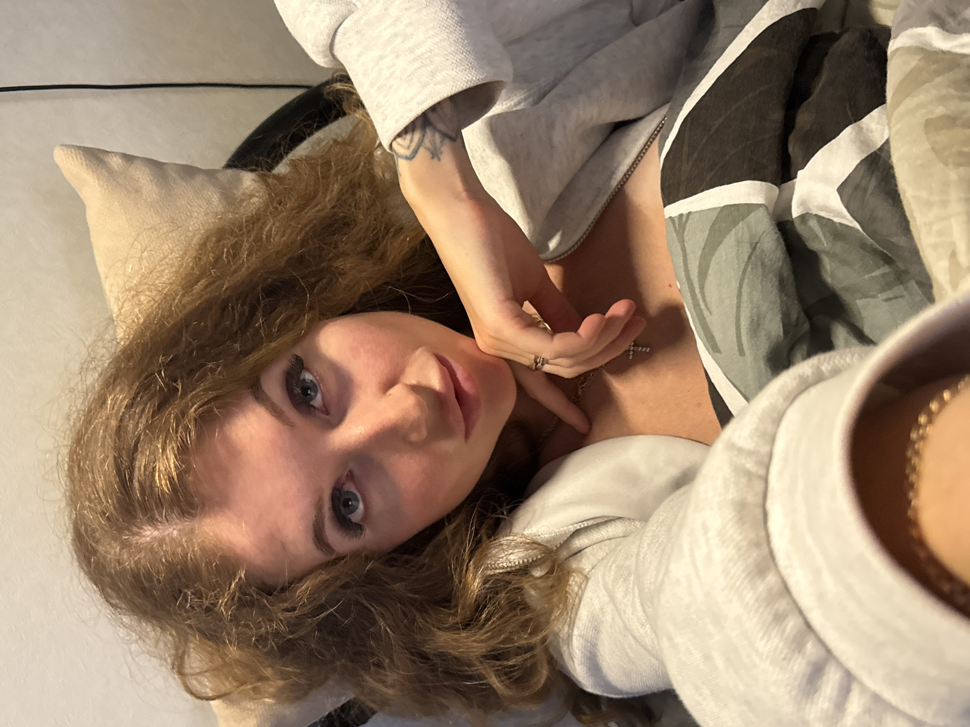 A woman lies on a bed. She has blue eyes, light skin, and curly brown hair. She is wearing a light grey sweatshirt and a cross necklace. Her right hand rests on her neck, fingers extended, and a ring on her ring finger. A pillow is located to her left. A black chair or object is located just above the pillow. A patterned duvet or blanket lies beside her. The location is undetermined, and it is not clear if the scene is in Chaikovsky, Russia.