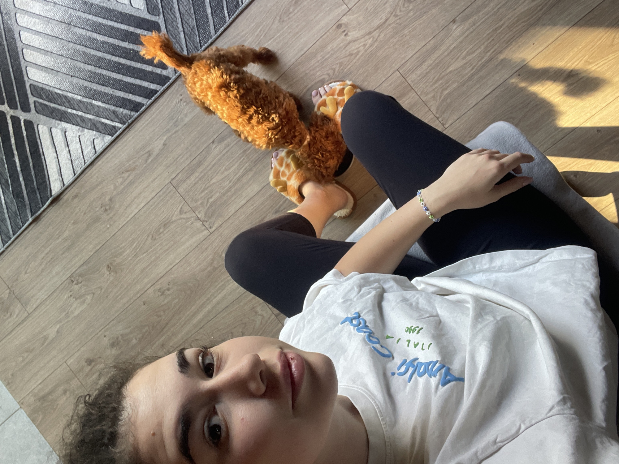 A woman wearing a white t-shirt with a blue and green logo and black leggings is lying on a gray seat, partially visible, in a room with light wood flooring. She has a beaded bracelet on her left wrist. Beside her on the floor is a tan poodle dog and giraffe-print slippers. A rug with a geometric design is in the upper left corner of the frame. Sunlight streams in from the right. The specific location in Sofia, Bulgaria, cannot be determined from the image.