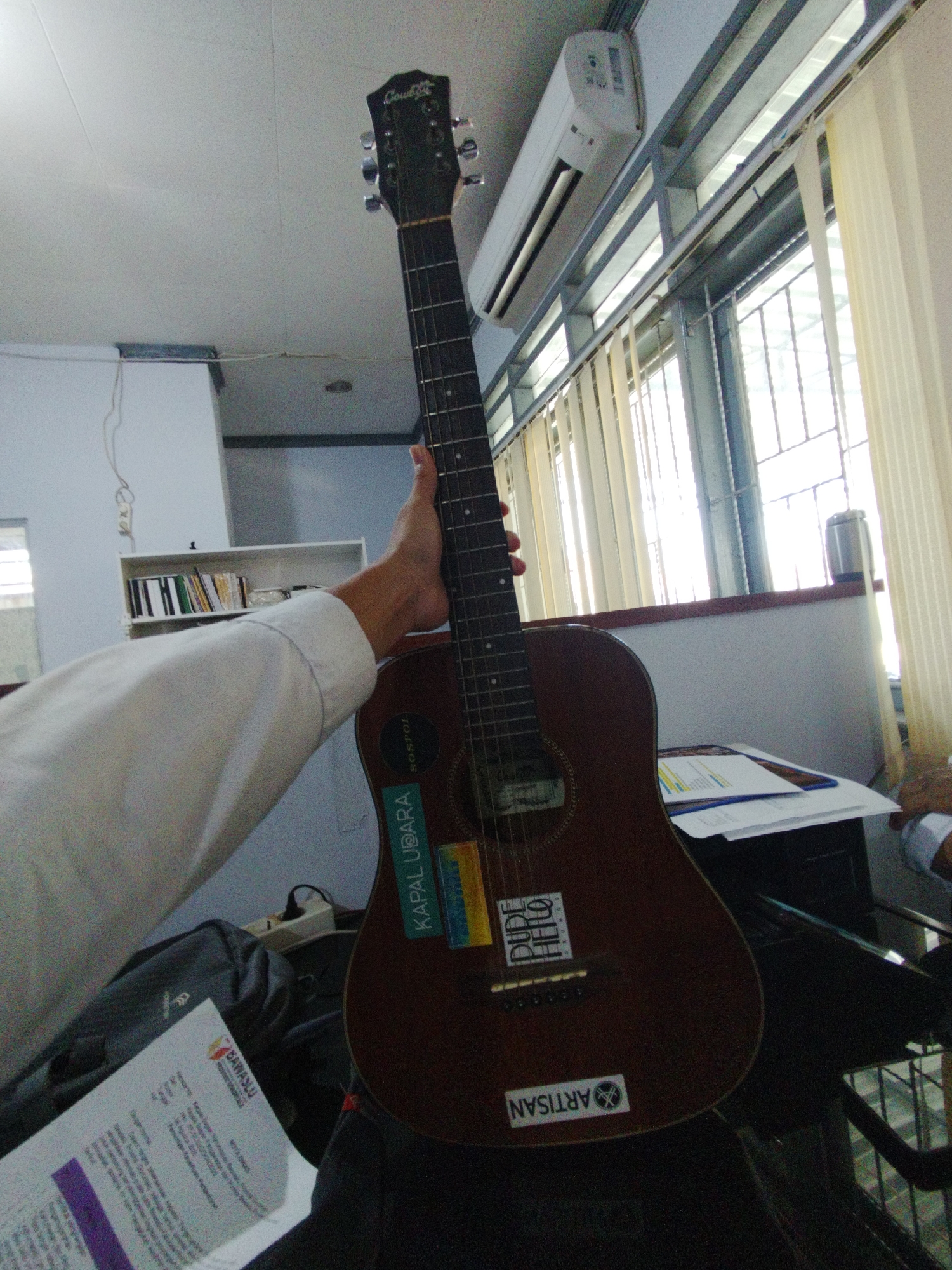 The image depicts an acoustic guitar being held by a person's arm, with the guitar's body and neck prominently featured. The guitar is dark brown and has several stickers on its body, including one that says "KAPAL UDARA" and another that says "ARTISAN". The person holding the guitar is wearing a white shirt. In the background, there is a shelf with books, a window with vertical blinds, an air conditioner mounted on the ceiling, and a desk with papers. The scene appears to be indoors, possibly an office or classroom, given the presence of the shelf, desk, and air conditioner. The lighting suggests it is daytime. There are no overt signs indicating this specific location within the city of Gorontalo, Indonesia, as the background elements are common to many indoor environments.