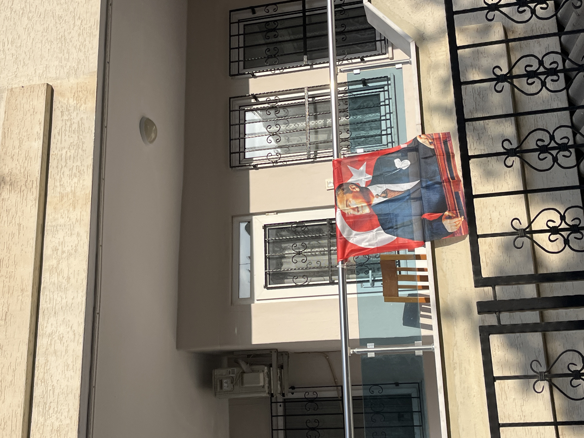 The image shows a building facade in Alanya, Turkey, with multiple floors. Windows are adorned with decorative iron grilles. A Turkish flag with an image of Mustafa Kemal Atatürk is attached to a pole in front of the building. Part of an iron fence with floral designs is visible to the right. An air conditioning unit is installed on the building.