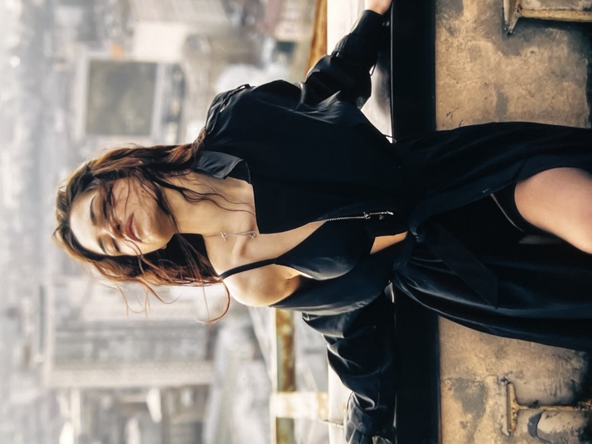 A woman with long, dark, wavy hair is positioned on a ledge or rooftop, facing away from the camera with her head tilted back and eyes closed. She is wearing a black bra and a black trench coat, which is draped off her shoulders. A silver necklace with a pendant hangs around her neck. Her arms are resting on the ledge, and the wind is blowing her hair. In the background, a blurred cityscape with buildings is visible under a cloudy sky. The setting appears to be urban, possibly on a rooftop overlooking a city.