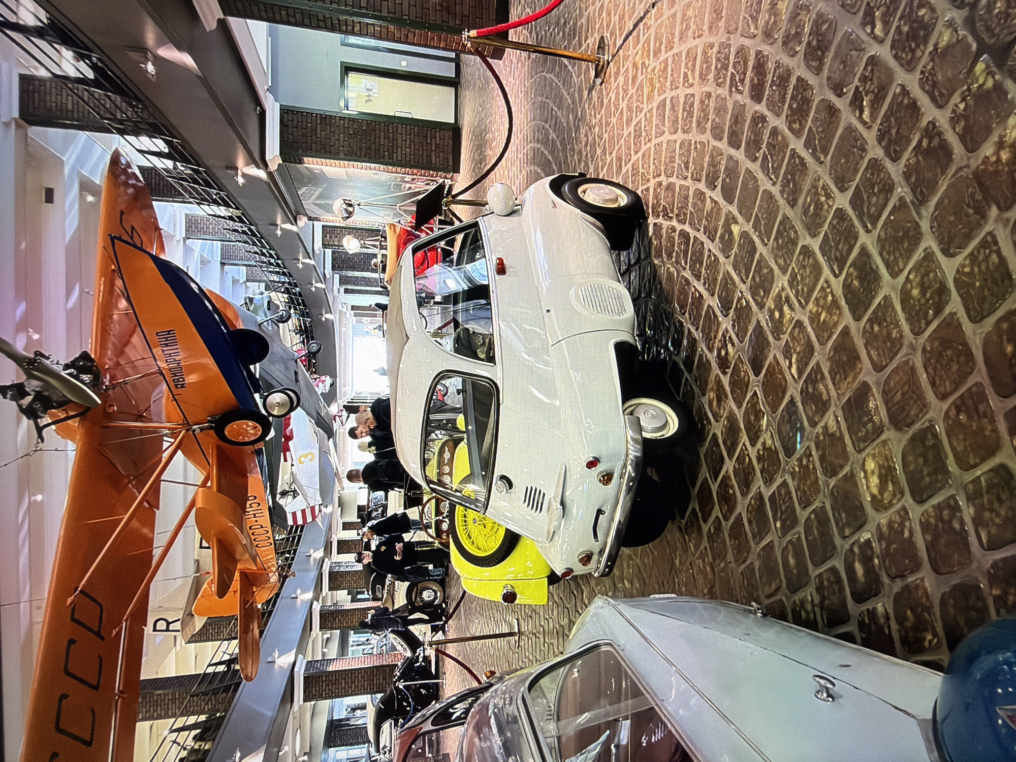 The image depicts the interior of a museum, likely a transportation museum, given the presence of vintage vehicles and aircraft. In the foreground, a white microcar, possibly a BMW Isetta, is prominently displayed on a cobblestone floor. To its left, a portion of another vintage car, light blue in color, is visible. In the background, several other cars are parked, and a yellow vehicle with a spare tire mounted on its rear is situated behind the white microcar.
Suspended from the ceiling are at least two vintage aircraft, one predominantly orange and the other white with red accents. The orange aircraft bears Cyrillic lettering, including "СССР" on its wing and "АВИАМАСТЕР" on its fuselage. The aircraft are suspended by wires and are positioned above the vehicles.
The museum's interior features brick pillars and balconies with railings, suggesting multiple levels or mezzanine areas. Several individuals are present in the mid-ground, observing the exhibits. One individual, dressed in what appears to be museum staff attire, is standing near the vehicles. Further back, a doorway with a "WC" sign is visible on the right.
The overall environment suggests an indoor exhibition space designed to showcase historical vehicles and aircraft. The lighting is a mix of ambient and artificial sources, casting shadows and highlighting the textures of the exhibits and the floor. The location is identified as a museum in Kotelniki, Russia.