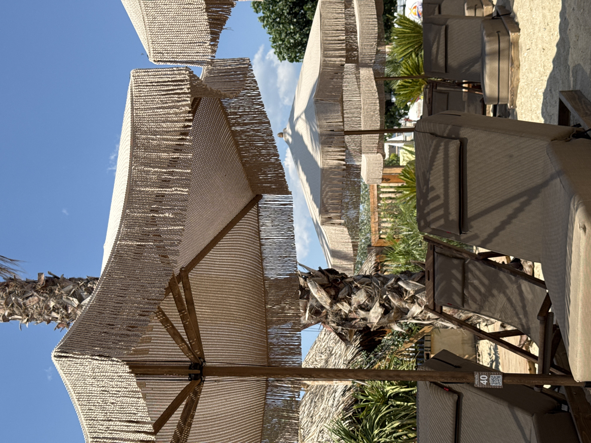 This image shows an outdoor scene at a resort or beach club, likely in a tropical or Mediterranean location. The primary subjects are lounge chairs and large, woven umbrellas providing shade. Several lounge chairs, upholstered in a neutral fabric, are arranged on a sandy or stone-paved ground. The umbrellas are made of a natural, woven material and feature fringe detailing, adding a bohemian or artisanal touch to the setting.
The scene appears to be set during the daytime, with bright sunlight casting shadows. The sky is a clear, vibrant blue with a few wispy clouds visible. Lush green palm trees and other tropical foliage are interspersed throughout the scene, reinforcing the vacation atmosphere. In the background, there are hints of other structures, possibly part of the resort, and more umbrellas and lounge chairs suggesting a well-equipped recreational area.
One notable detail is a small sign attached to a wooden support beam of one of the lounge chairs. It displays the number "40" prominently, along with a QR code. This could indicate a specific area, reserved seating, or a service offering. The overall impression is one of relaxation and leisure, with the setting designed for comfort and enjoyment in a sunny, warm environment. There are no people visible in the image, focusing the attention on the tranquil setup.