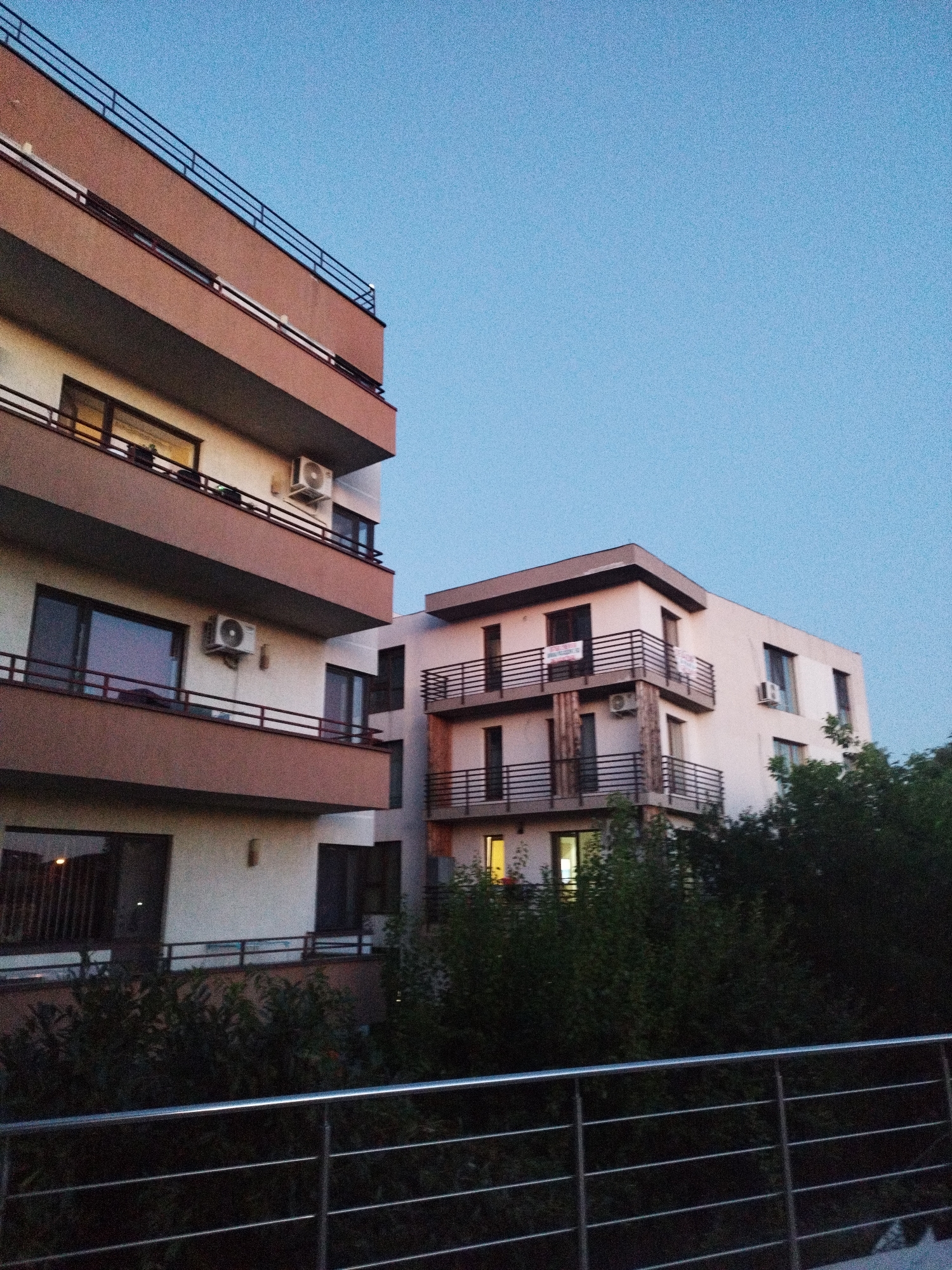 The image depicts two apartment buildings in Bucharest, Romania, as evening light fades. The building on the left is a modern, multi-story structure with protruding balconies, each bordered by dark railings. The facade features a reddish-brown trim against a white background. Air conditioning units are visible on several floors. The building on the right is also a multi-story apartment building, with a lighter colored facade and similar balconies with dark railings. One of these buildings has a sign hanging on a balcony, likely advertising an apartment for sale or rent. Dense green foliage, representing trees and bushes, partially obscures the lower sections of both buildings. In the foreground, a silver metal railing runs horizontally across the frame. The sky above is a clear, deep blue, characteristic of twilight. There are no people or any specific actions or interactions identifiable in the image.