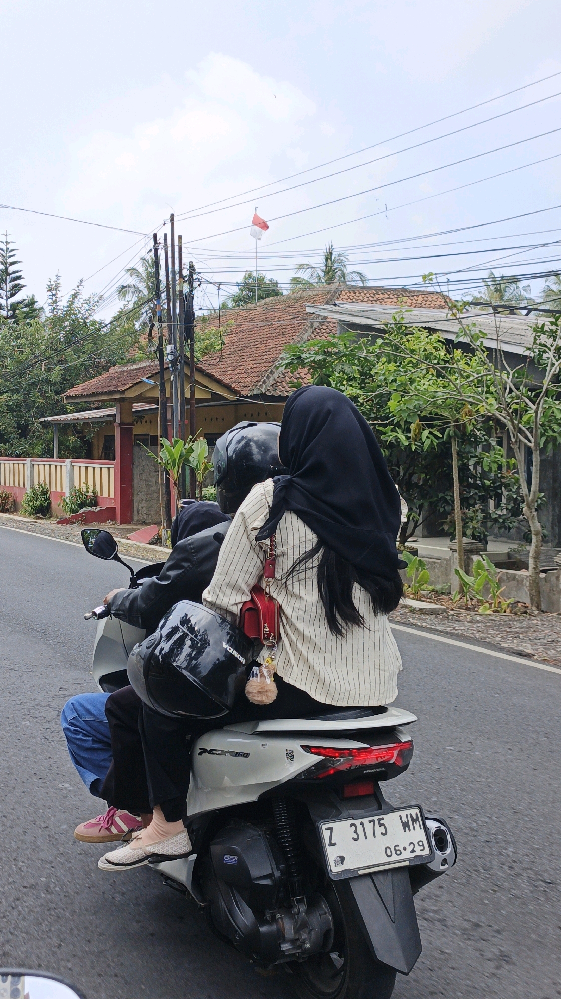 Two individuals are riding on a white scooter on a paved road in Sukaresik, Indonesia. The rider in the front is wearing a black helmet, a dark jacket, and dark pants. The passenger in the back is wearing a black hijab, a light-colored striped shirt, and dark pants. They are both facing forward, and the passenger is holding a red bag with a keychain attached to it. A black helmet is placed on the rear of the scooter. The license plate of the scooter reads "Z 3175 WM 06-29". The environment includes houses with terracotta roofs, trees, and utility poles with overhead wires. The sky is overcast.